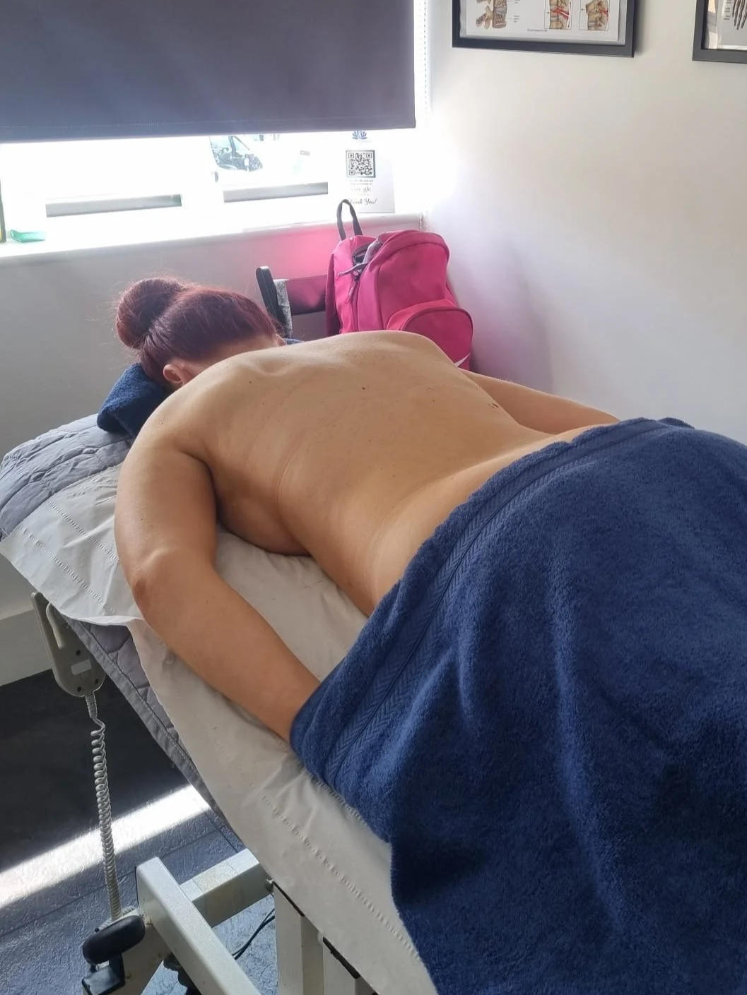 A woman lying face down on a massage or therapy table in a clinical setting, with her head turned to the side. The room has framed diagrams on the wall, a window with sunlight, and a pink backpack on a chair.