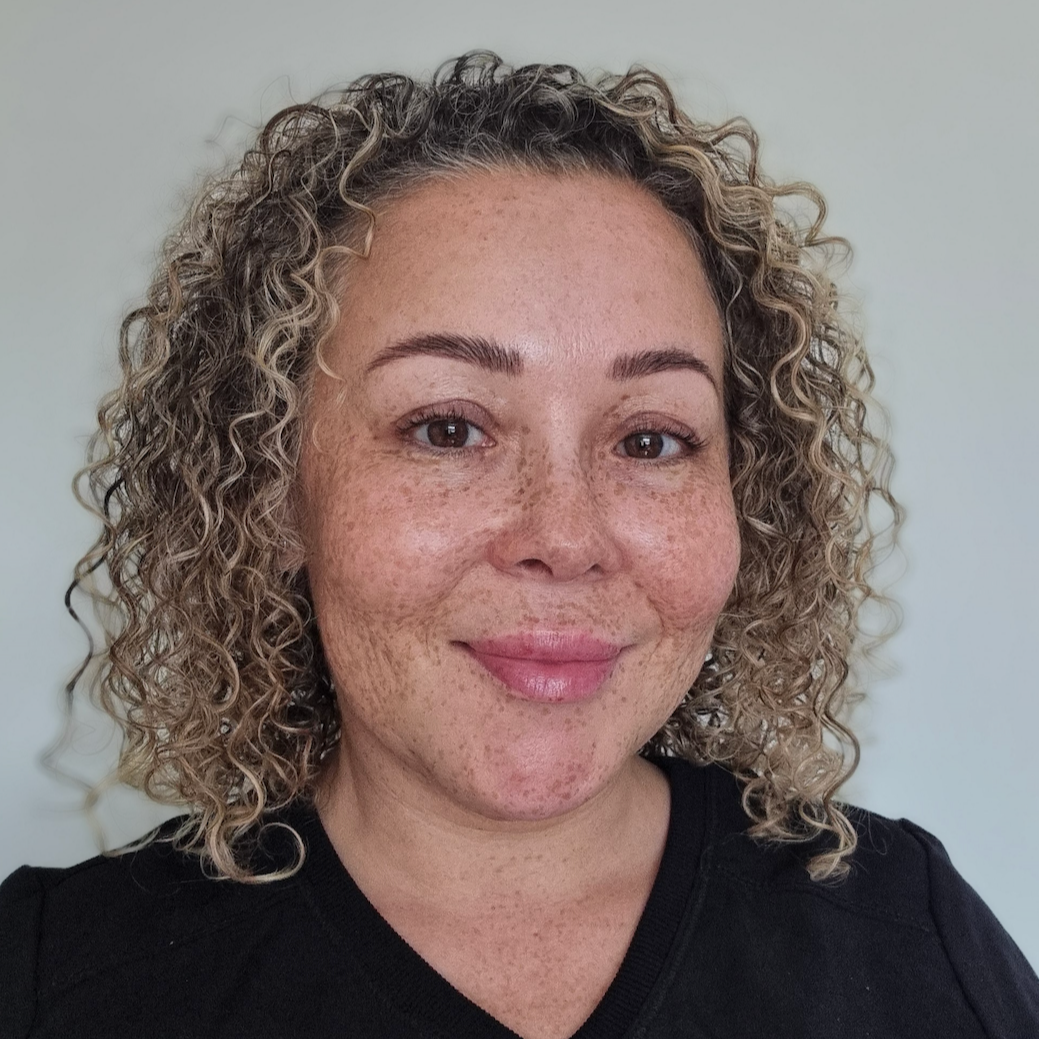 A woman with curly blonde hair smiling in front of a plain light-colored wall.