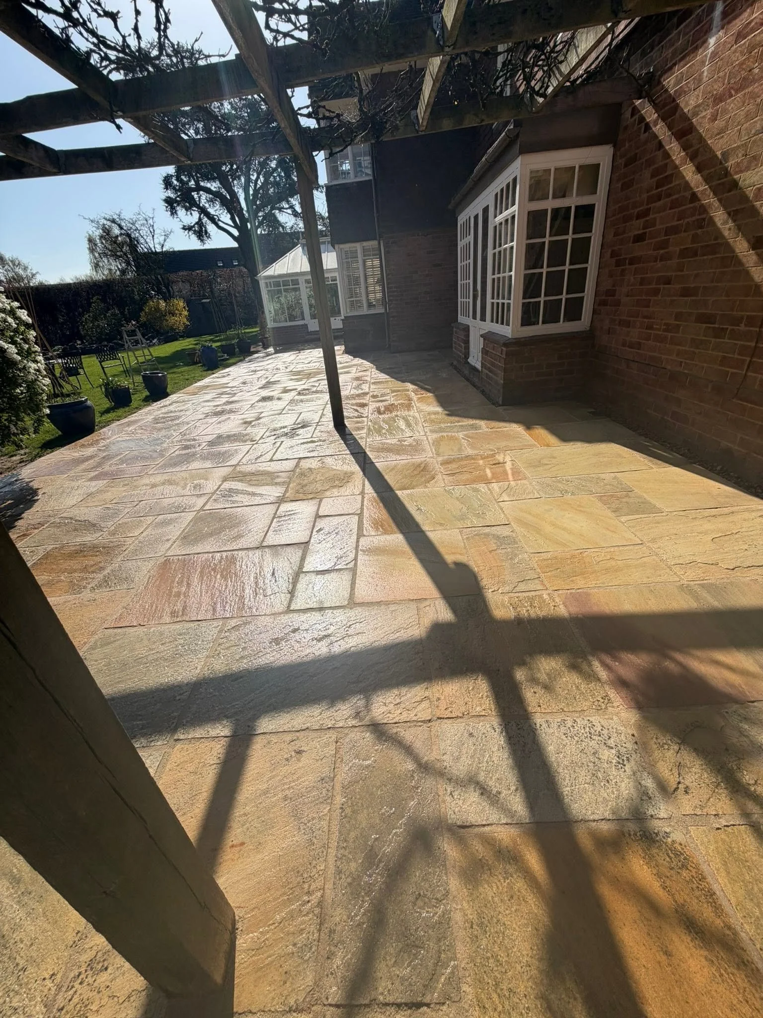 A paved patio area with several potted plants, a wooden pergola, and a garden in the background with trees and bushes. The photo is taken on a sunny day, casting shadows on the ground.