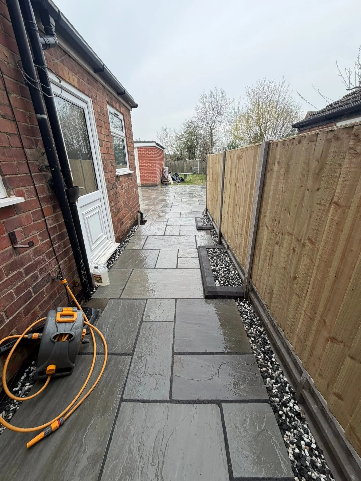 A backyard patio with newly installed large gray stone tiles, black and white pebble border, wooden fence on the right, brick house on the left, and some tools and equipment visible in the scene.
