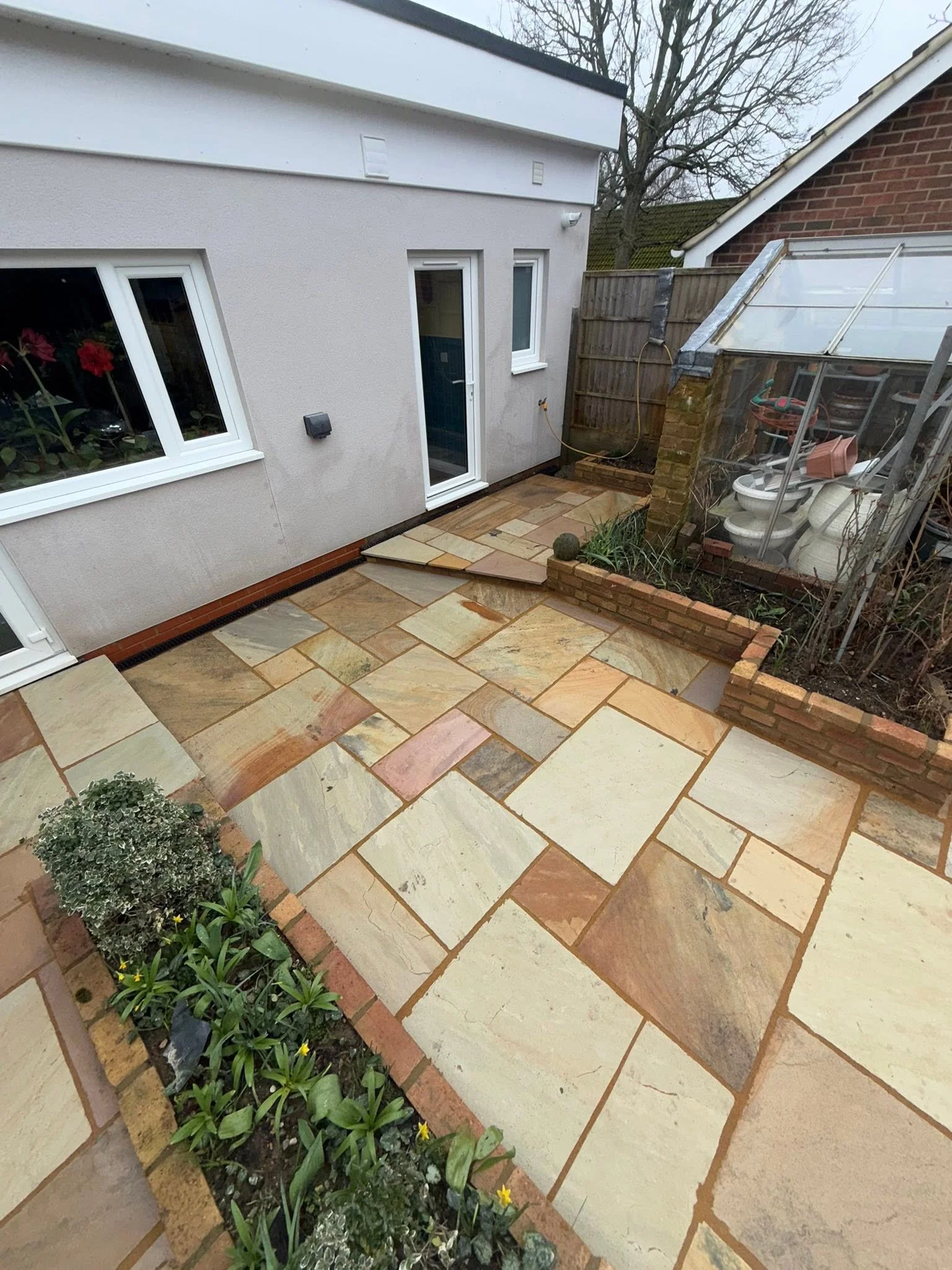 Small garden with an assortment of green plants and yellow flowers along a brick-edged border, with a patio made of large rectangular sandstone tiles and part of a house with a white exterior wall, a window, and a door. A wooden fence and a greenhous