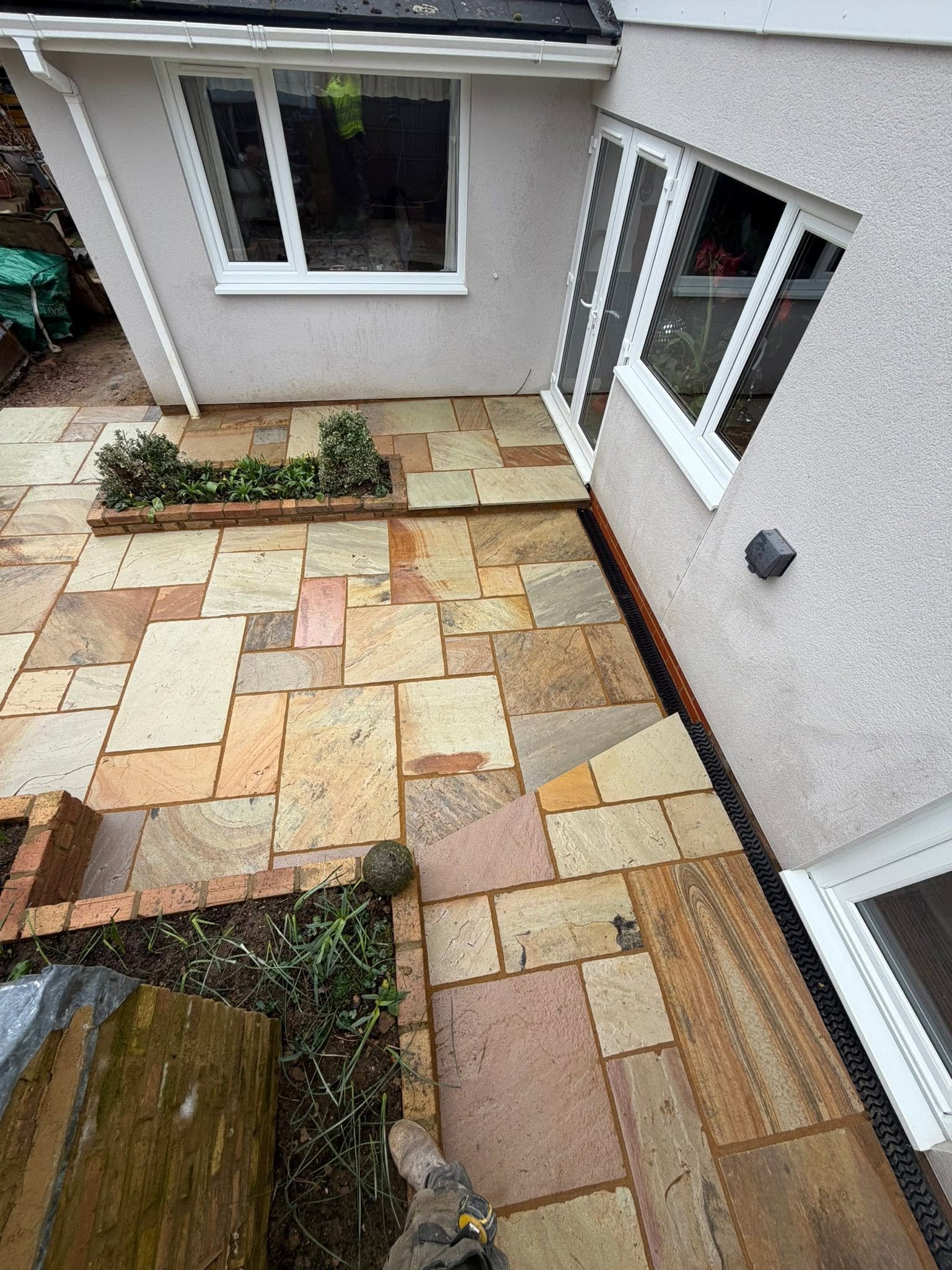 A backyard patio with multi-colored stone tiles, a small garden bed with plants, and an outside wall with two large windows and a door.