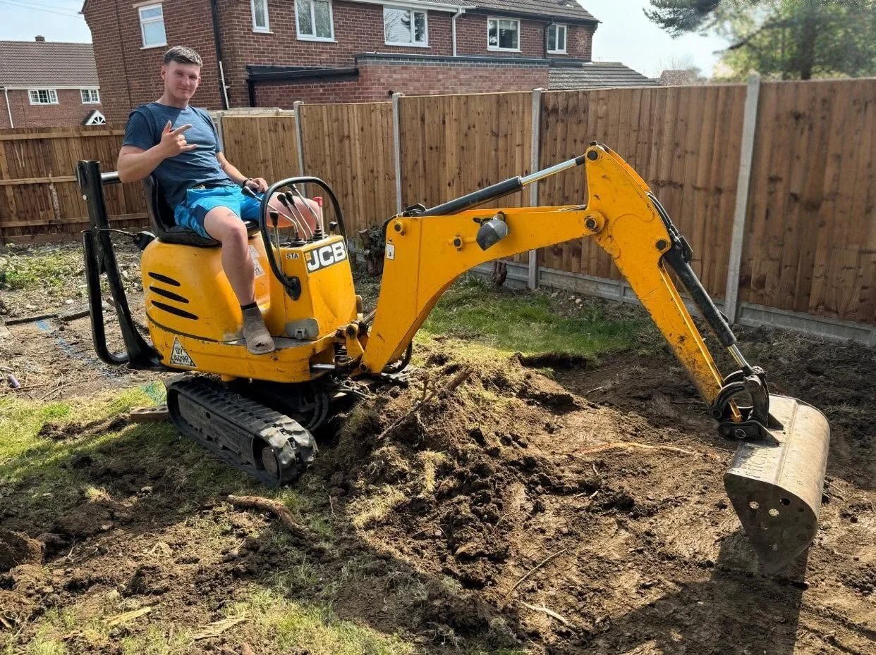 A young boy sitting on a small yellow JCB mini excavator in a backyard, giving a peace sign, with a wooden fence and brick houses in the background.