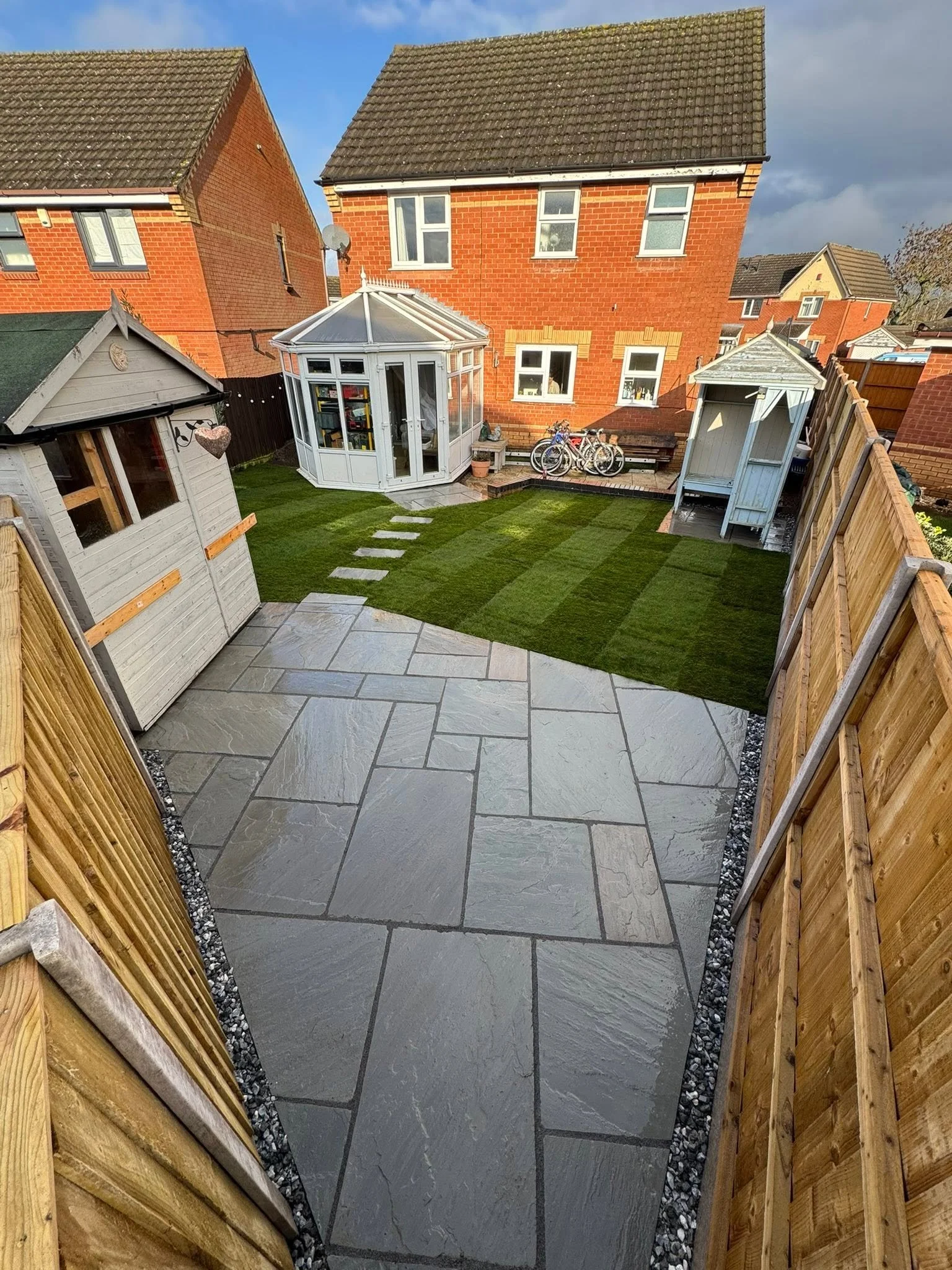 Backyard with artificial grass, stepping stones, wooden fence, small wooden shed, white greenhouse, and brick house in the background.