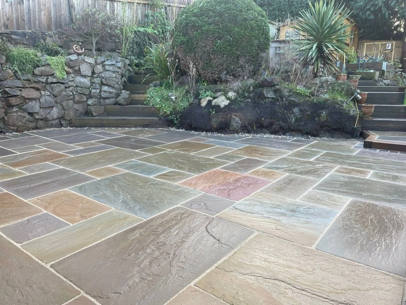 Backyard patio with large, multicolored stone tiles, stone retaining wall, garden, and stairs leading up to a garden area with plants and potted plants.