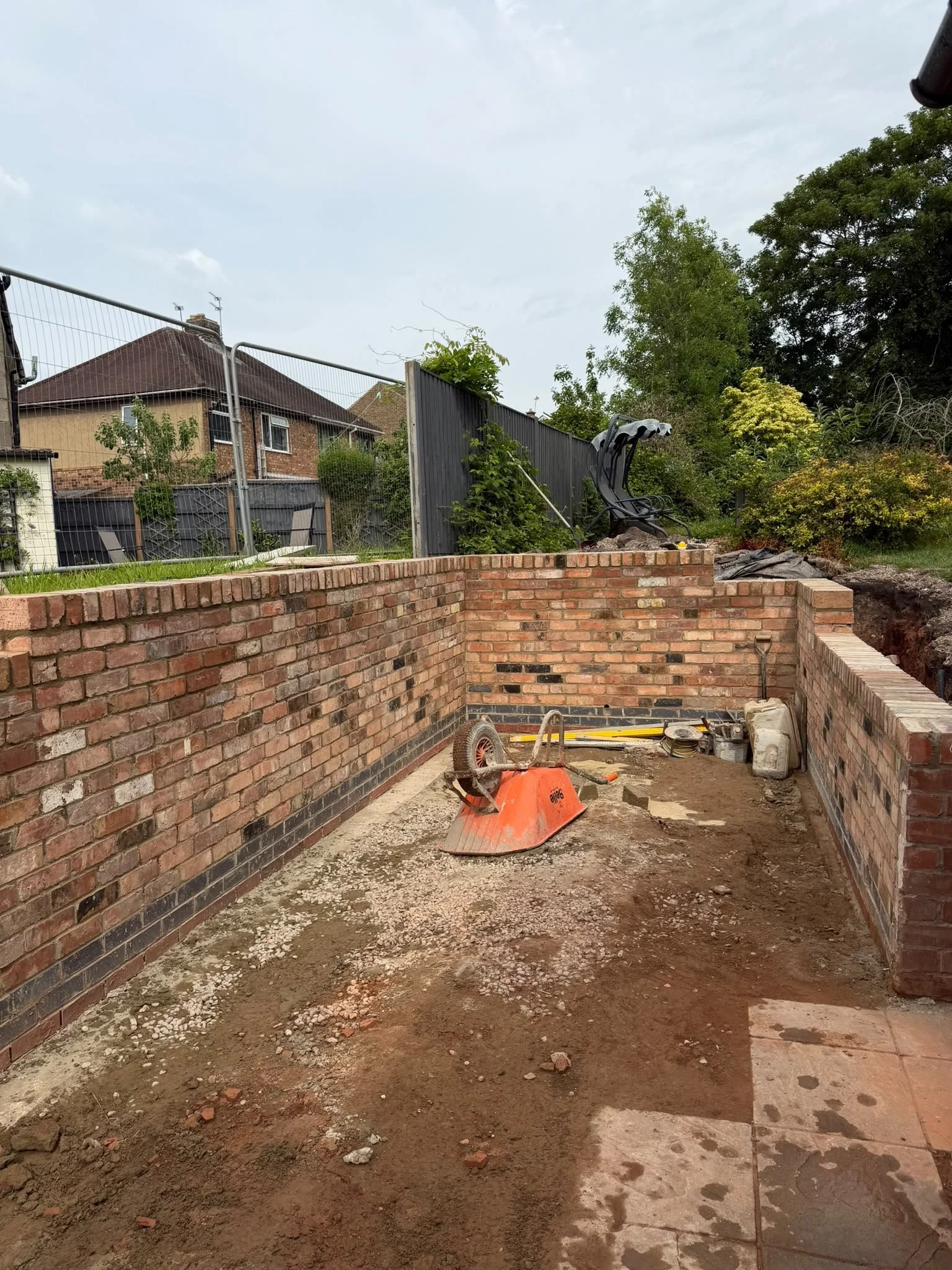 Construction site in a backyard with brick walls, a wheelbarrow, construction tools, and dirt ground, surrounded by trees and houses.