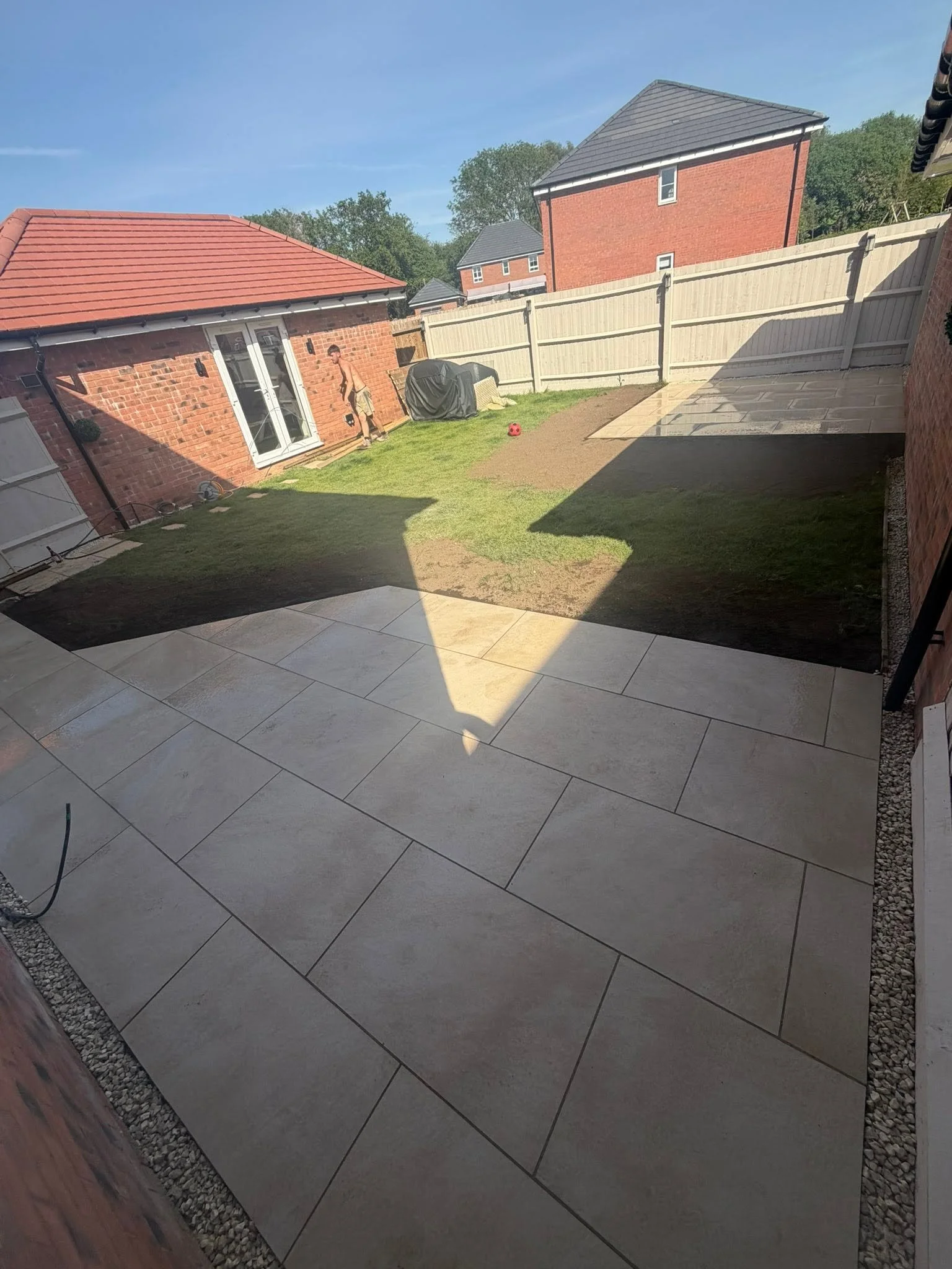 Backyard patio area with tiled flooring, a small grass patch, a shed, a fence, and neighboring houses under a clear blue sky.