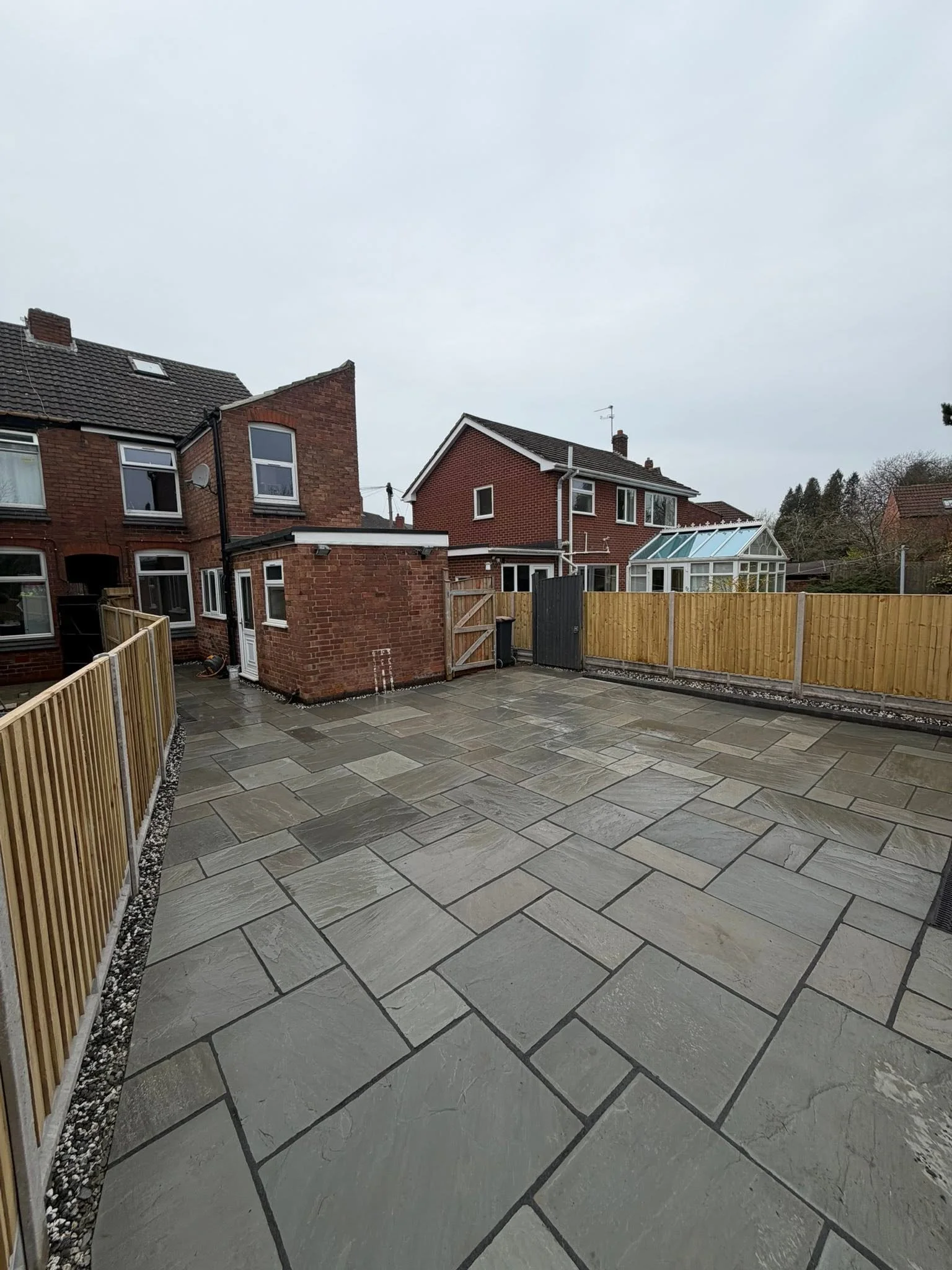 View of a residential backyard with a paved stone patio, wooden fencing on the left and right, brick houses with pitched roofs and a glass conservatory in the background, overcast sky.