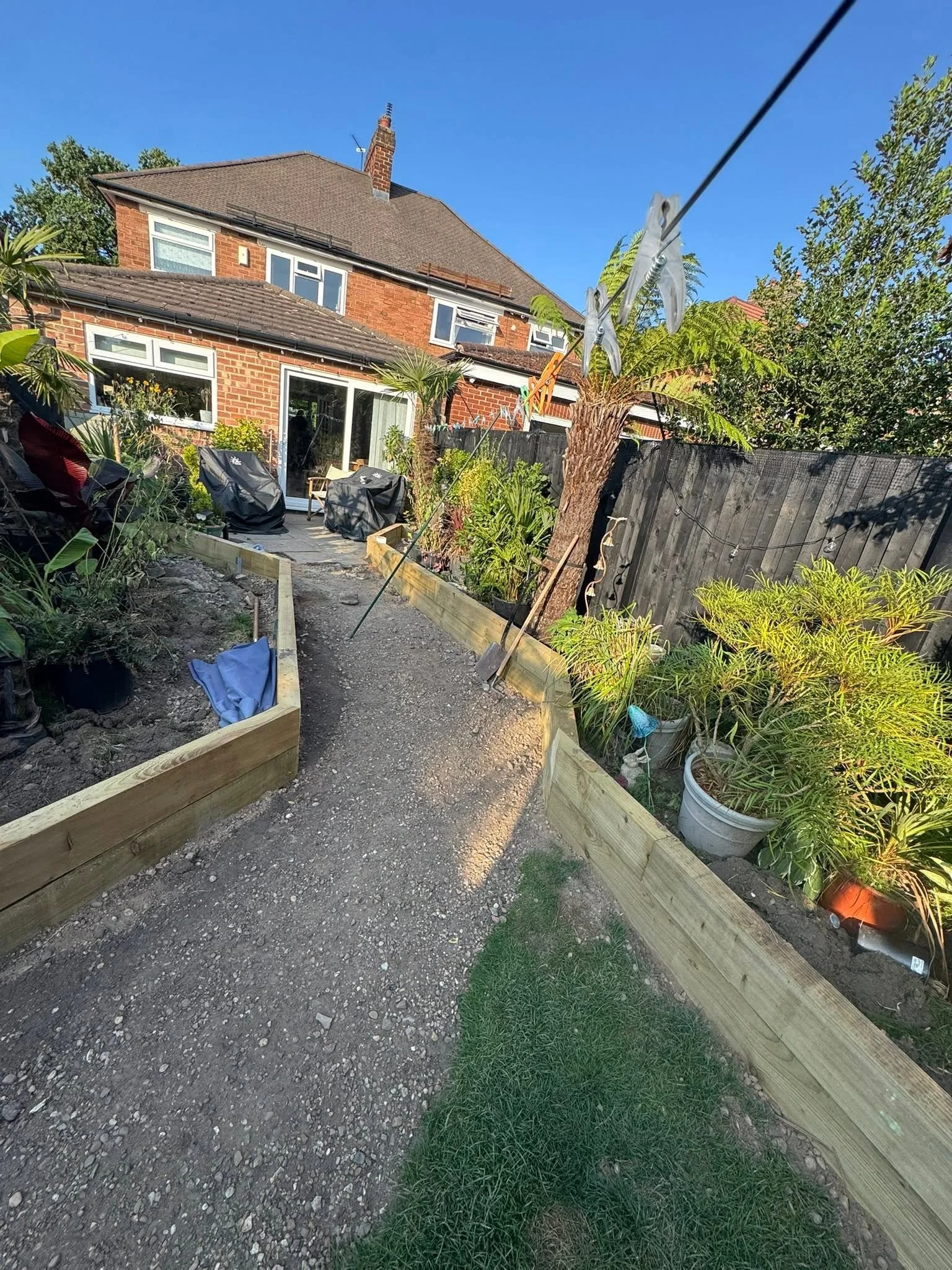 A backyard garden with new wooden garden beds, potted plants, and a string of outdoor lights. A house with brick walls and large windows is in the background, with a clear blue sky above.