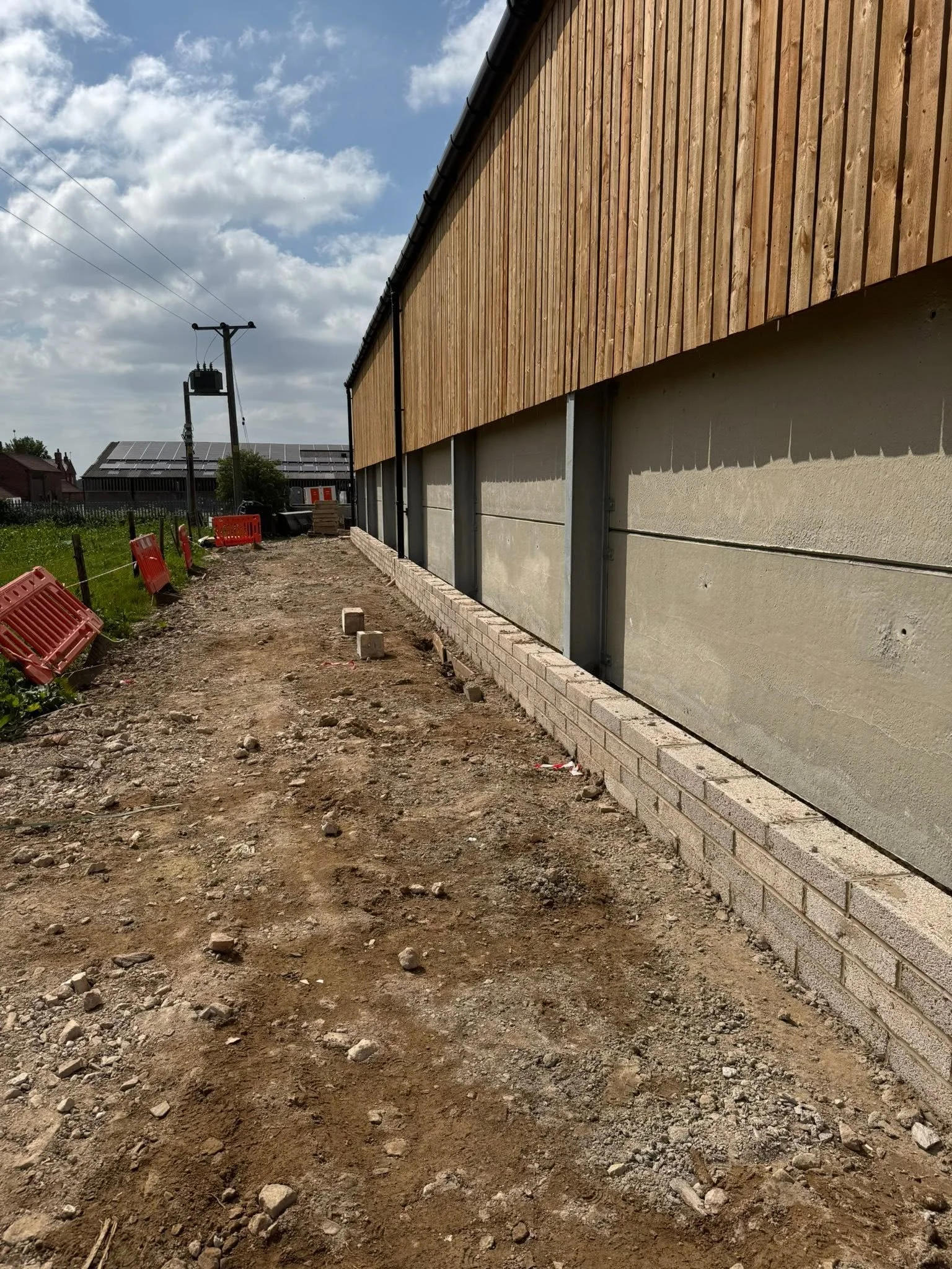 Construction site with a dirt pathway, wooden fencing on the right, and utility pole with power lines against a partly cloudy sky.