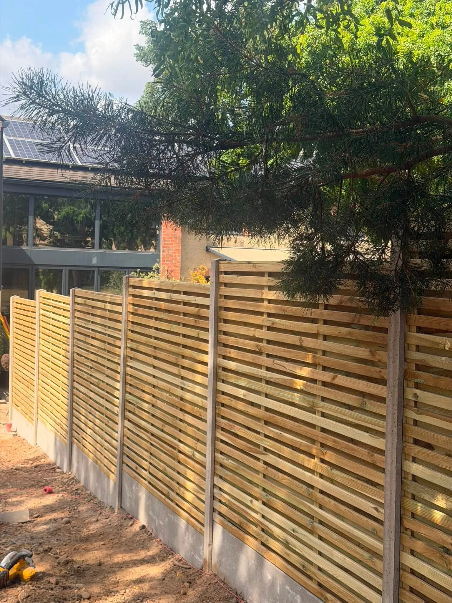 A newly constructed wooden fence with horizontal slats sits along a concrete foundation in a backyard. Behind the fence, there are trees and a house with solar panels on the roof, visible through a window. The ground in the foreground is dirt, with s