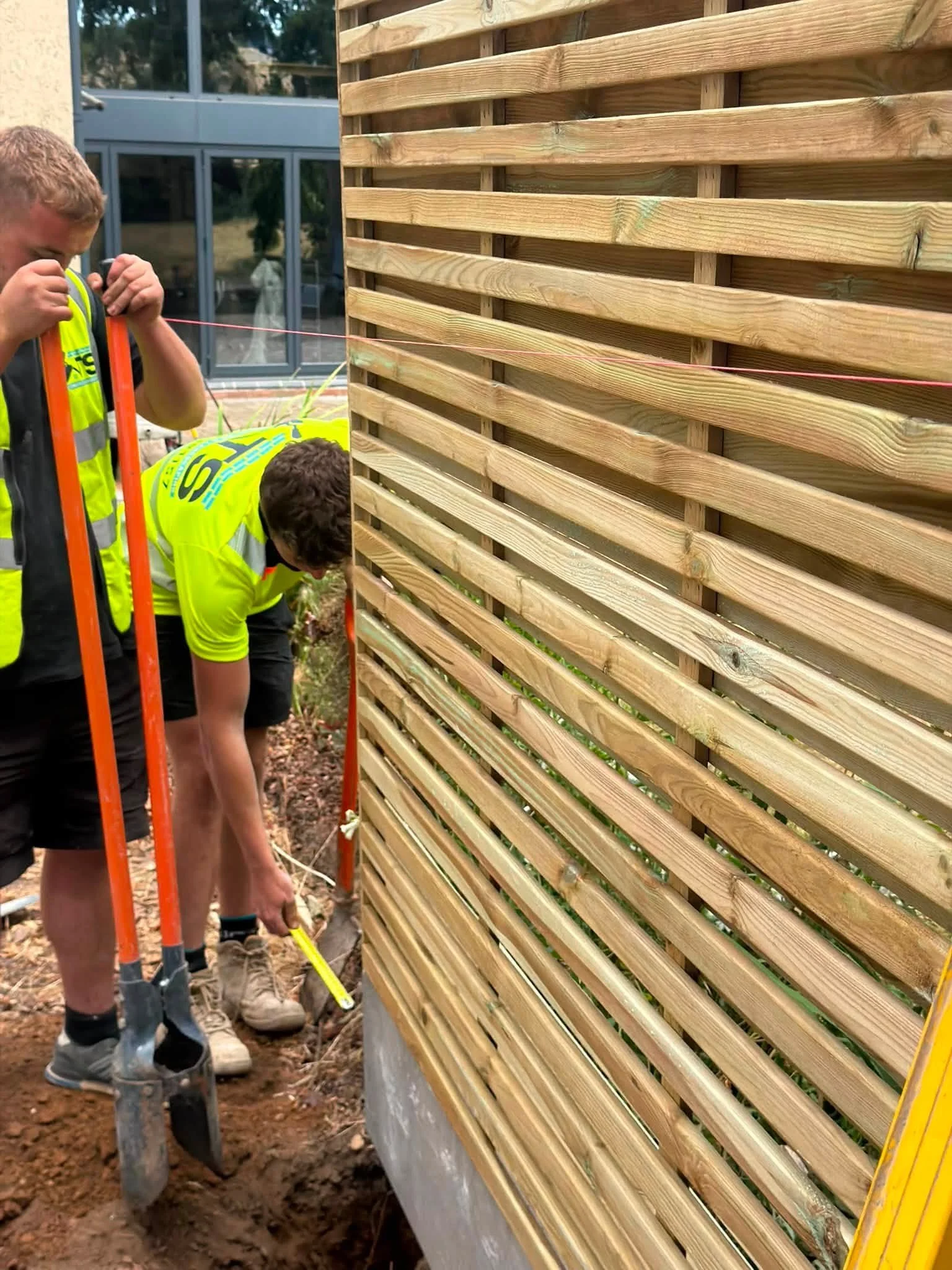 Two workers in yellow safety vests working on a construction project next to a wooden fence with metal supports, with a glass building in the background.