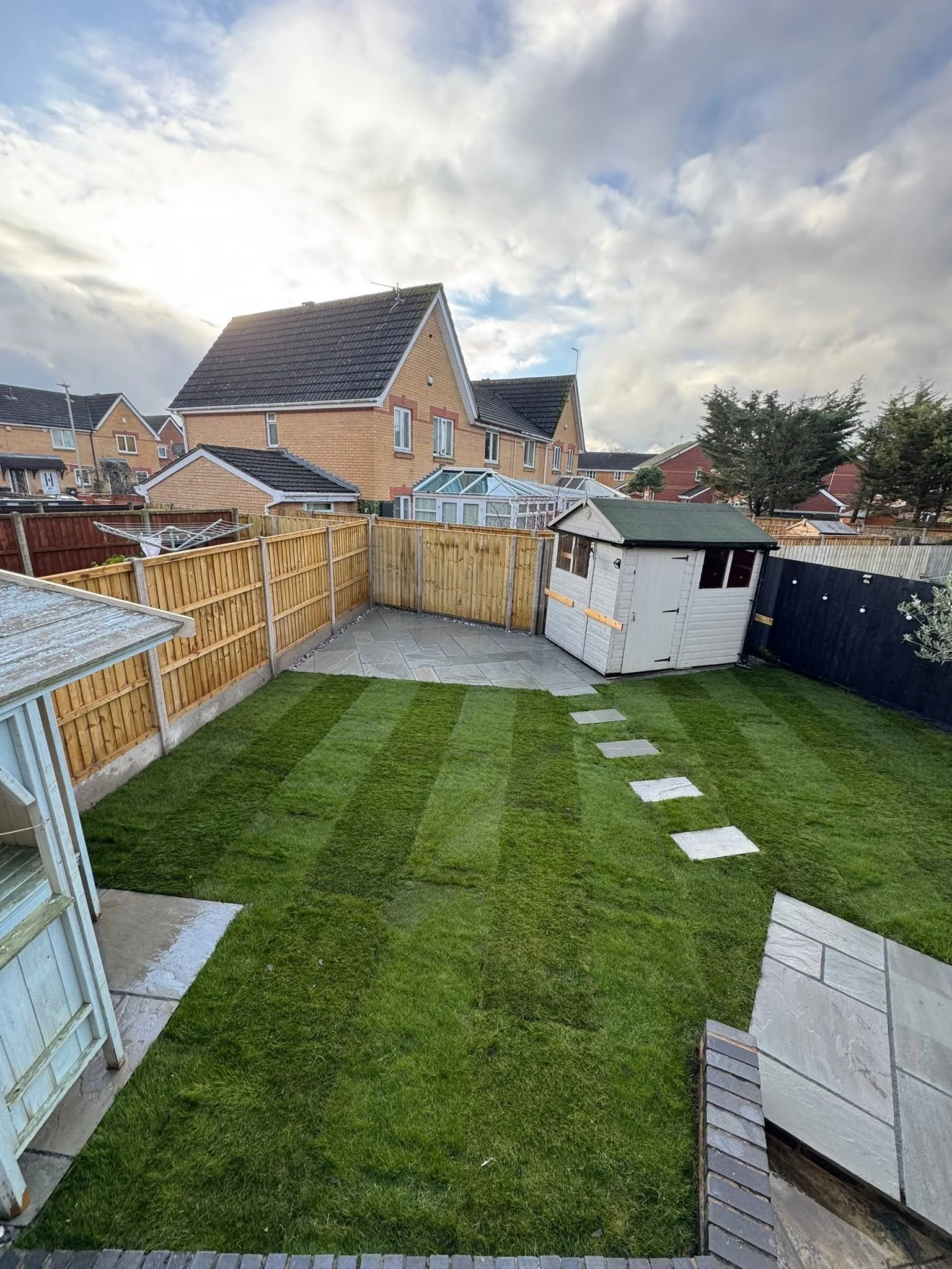 View of a backyard with a well-maintained grassy lawn, outdoor paving stones forming a path, a small white garden shed, and a wooden fence enclosing the yard. In the background, there are neighboring houses with brick exteriors and pitched roofs, som