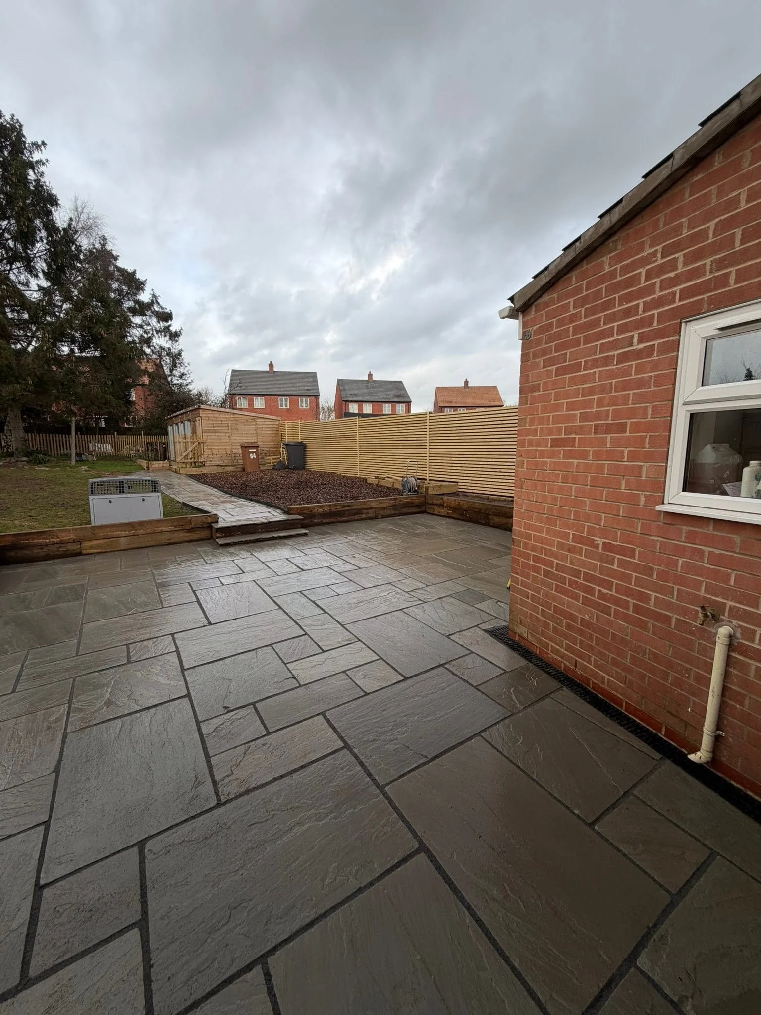 A backyard patio with gray flagstone tiles, a wooden raised garden bed, a brown trash bin, a gray compost bin, a wooden fence, and houses with pitched roofs in the background under a cloudy sky.