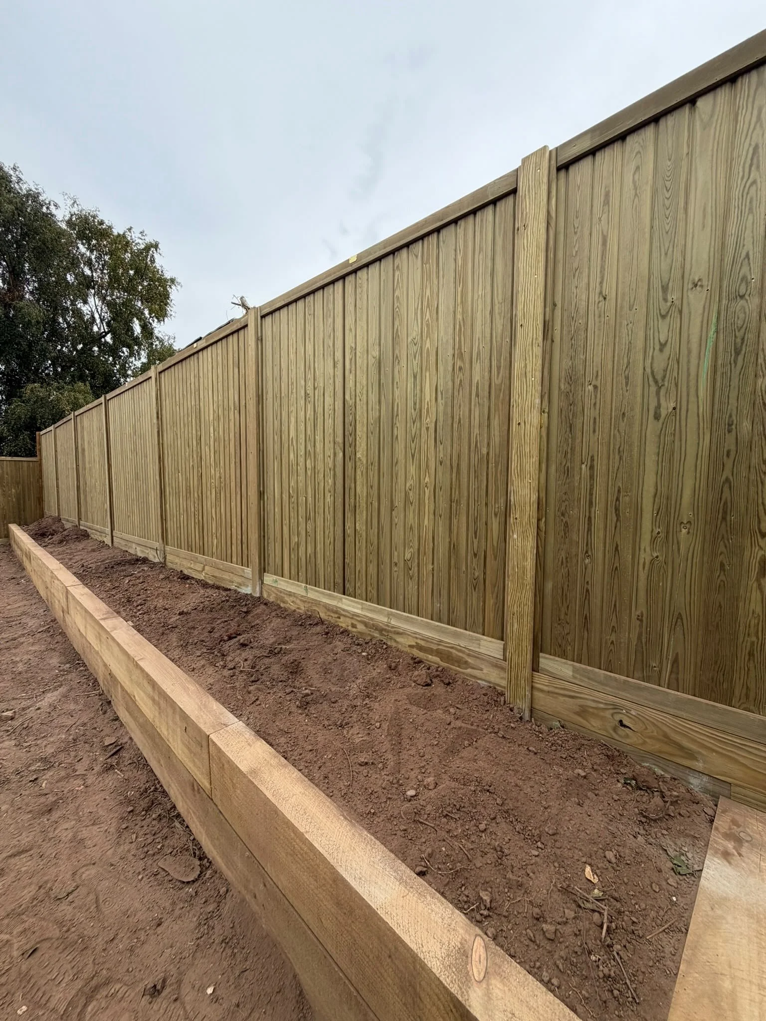 A wooden privacy fence along a dirt soil garden bed with a raised wooden border.