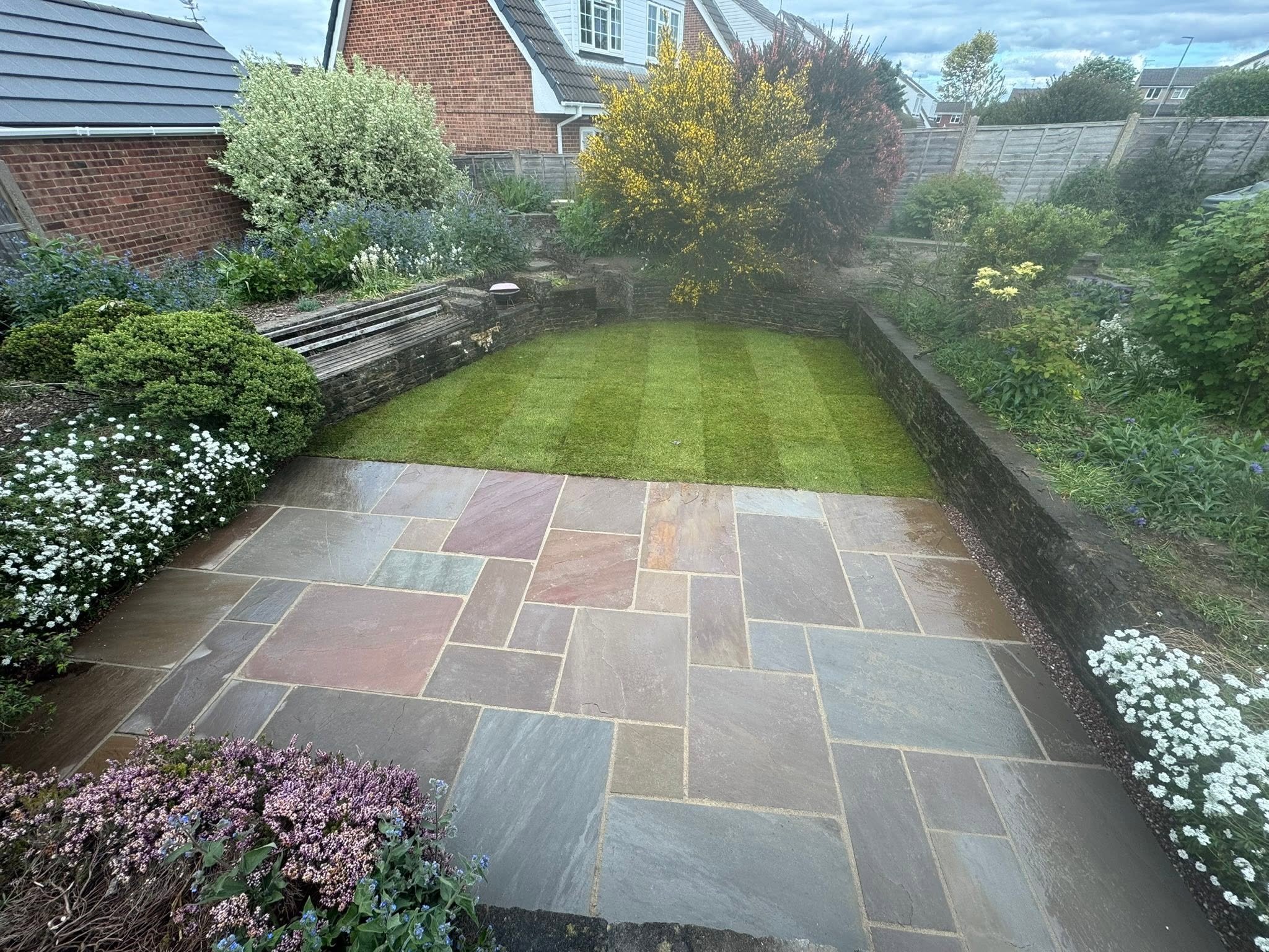 A landscaped backyard with a small, well-maintained lawn, bordered by flower beds containing blooming plants and shrubs. The yard is enclosed by a wooden fence and features a paved patio area with colorful stone tiles.