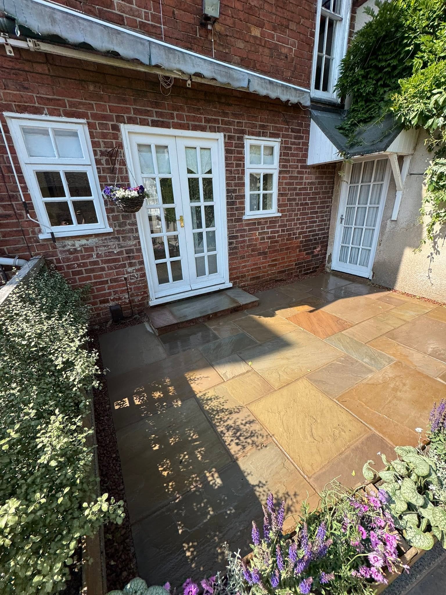 Backyard patio with stone flooring, brick house wall with white-framed glass doors, window, hanging flower basket, lush green plants, and shaded areas.