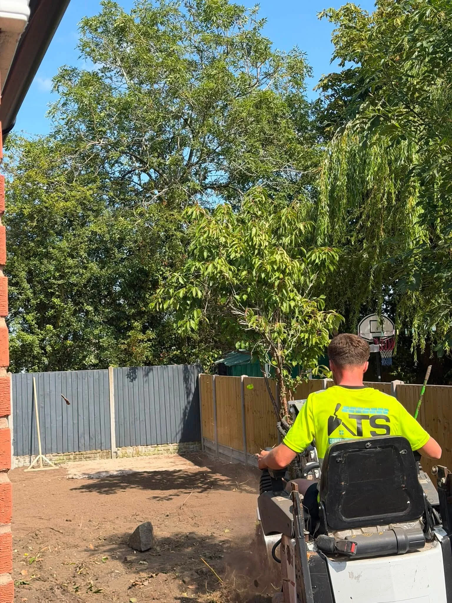 A person in a yellow construction shirt is operating a riding lawn mower or similar equipment in a backyard. There is a newly built wooden fence, trees, and a basketball hoop in the background.