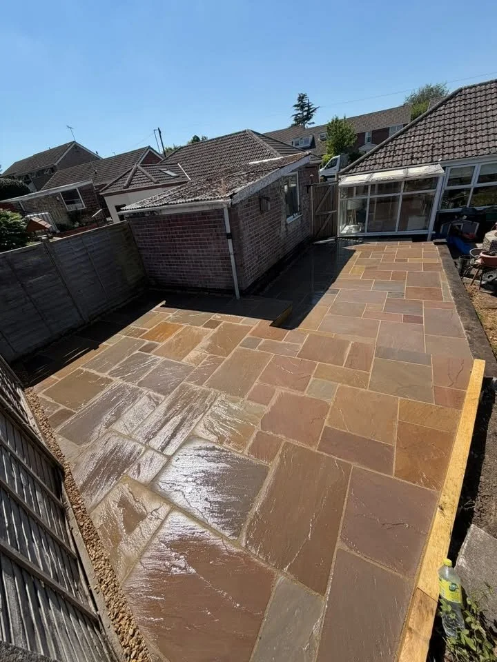 Newly laid stone patio in a backyard, partially wet from recent cleaning or sealing, surrounded by a wooden fence and neighboring houses with brick and tiled roofs.
