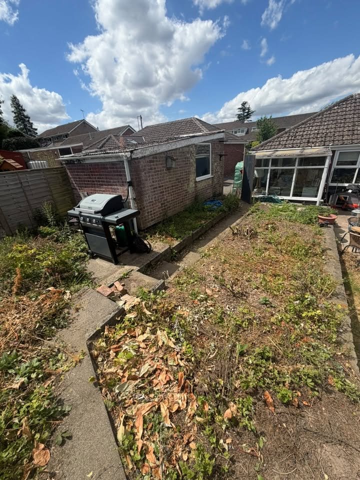 Backyard with a barbecue grill, overgrown plants, a brick building, and a small screened-in porch under a partly cloudy sky.