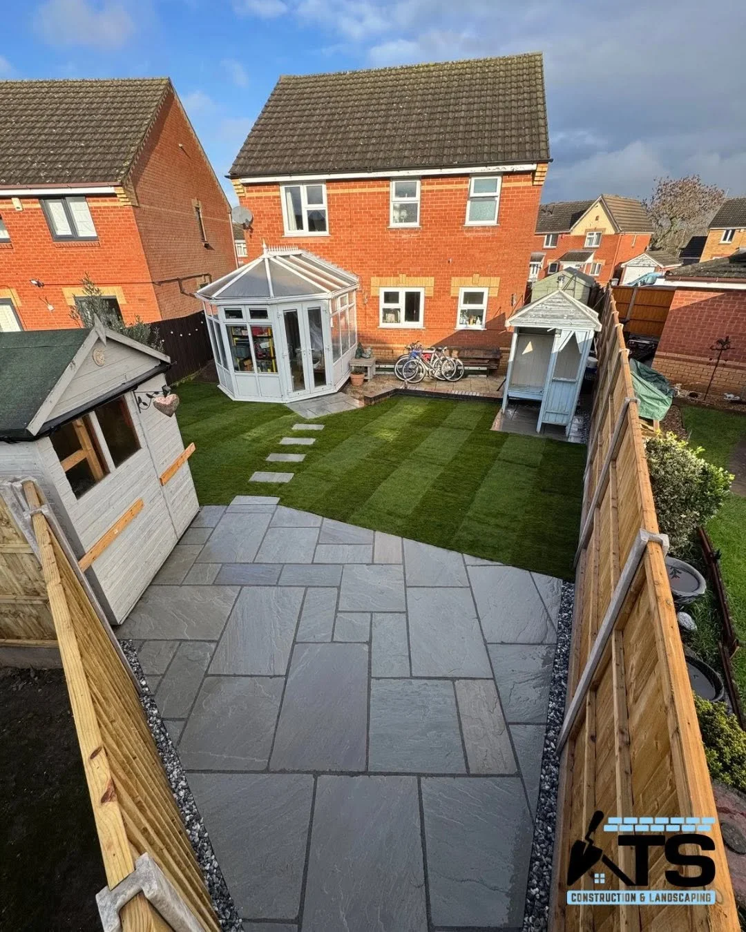Backyard with stone patio, striped lawn, and a wooden fence. Features include a small shed, a greenhouse, and a garden shed, with houses visible in the background.