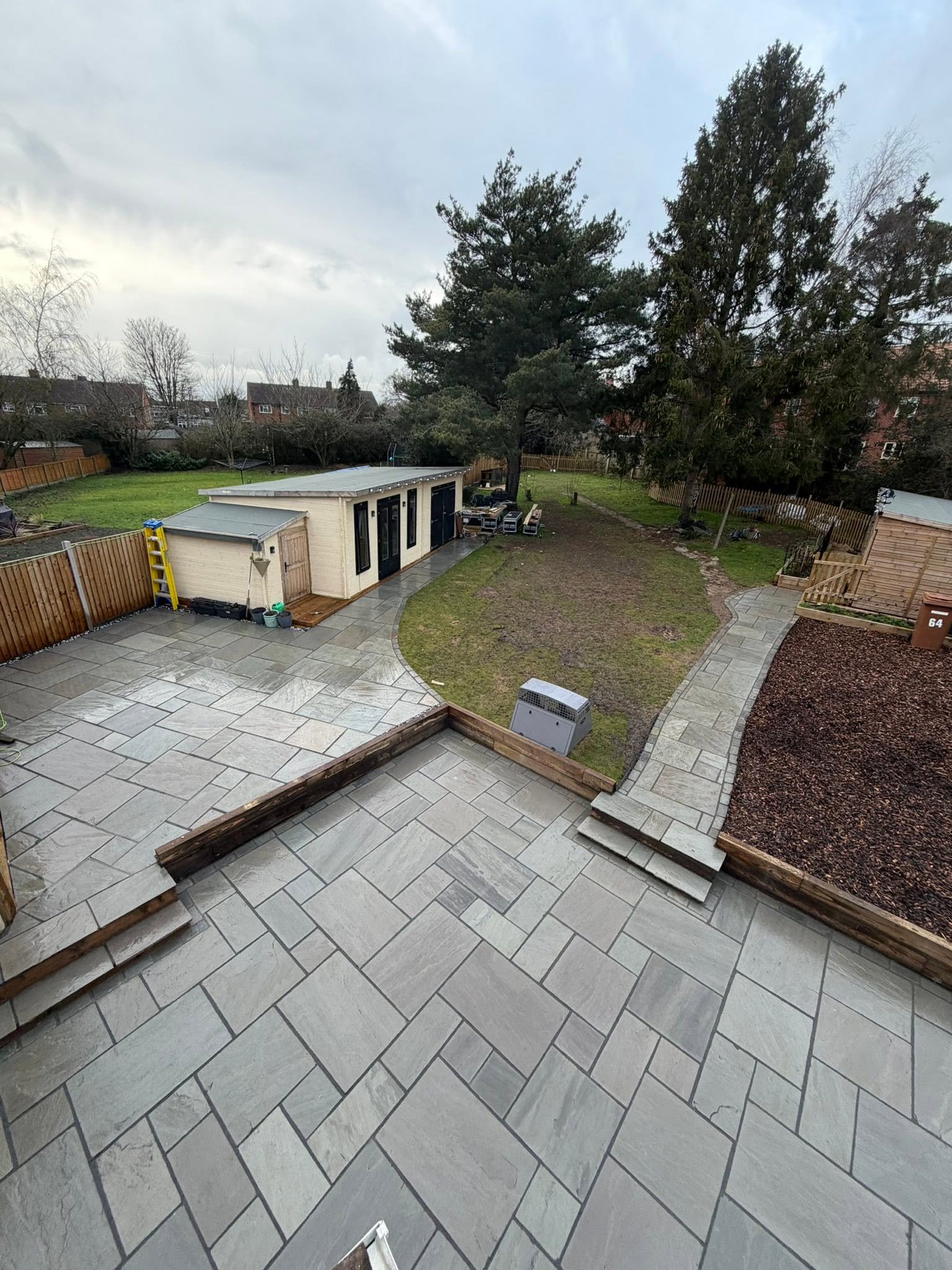 Backyard with a patio of large gray tiles, a small white building with black doors, a grassy area, and tall trees. There are construction supplies and tools near the building.