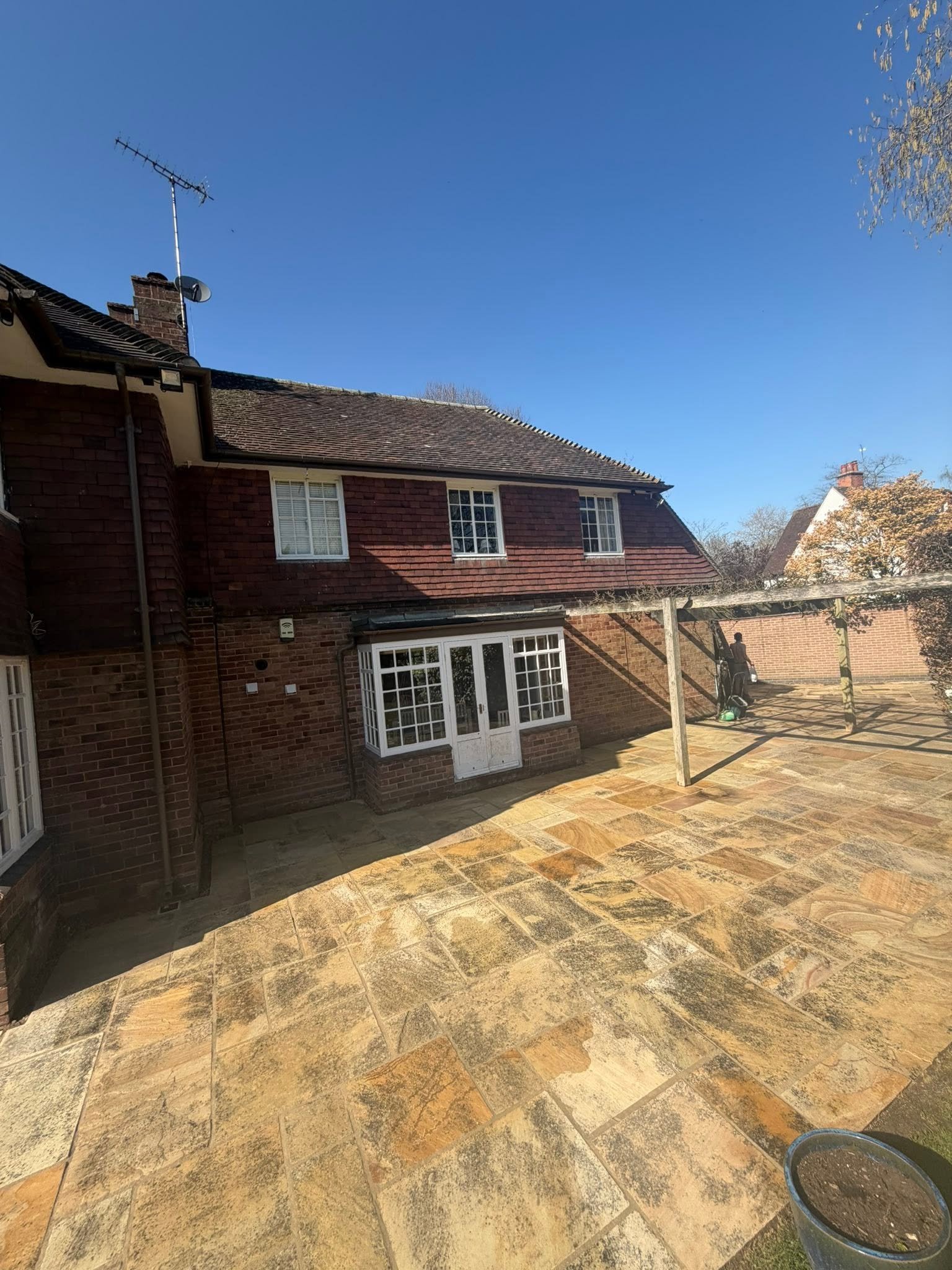 Backyard patio with stone tiles, a brick house with white-framed windows and a glass door, a wooden pergola, a person gardening, and a clear blue sky.