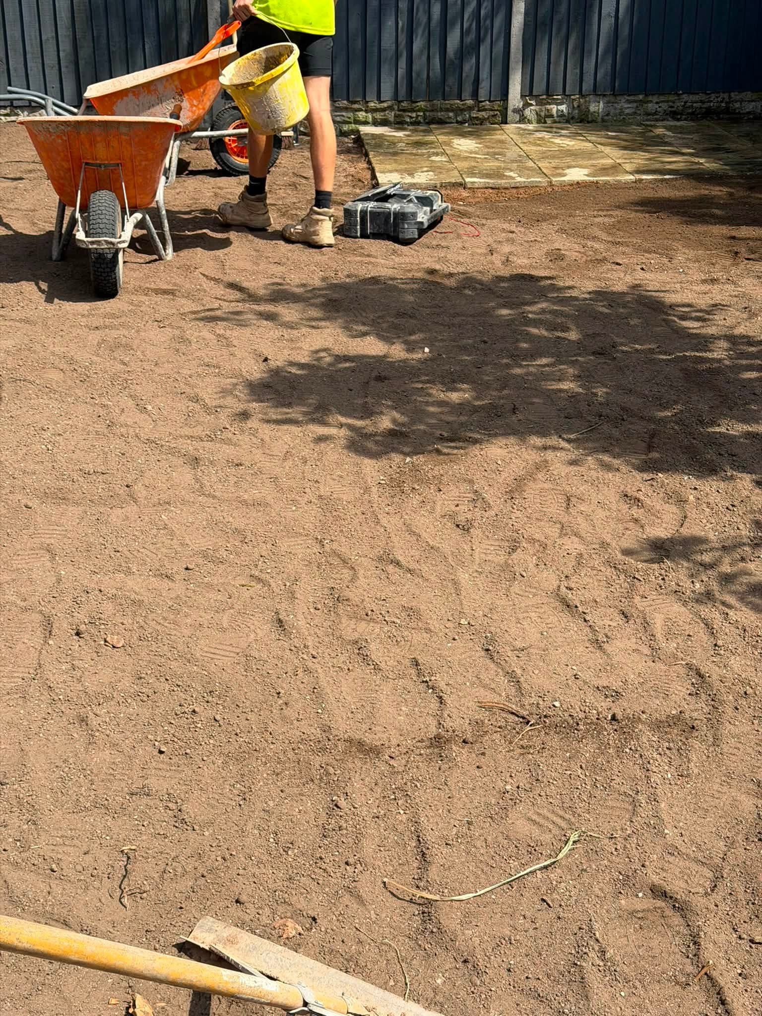 A person working on a dirt yard with a wheelbarrow, a toolbox, and paving stones in the background.