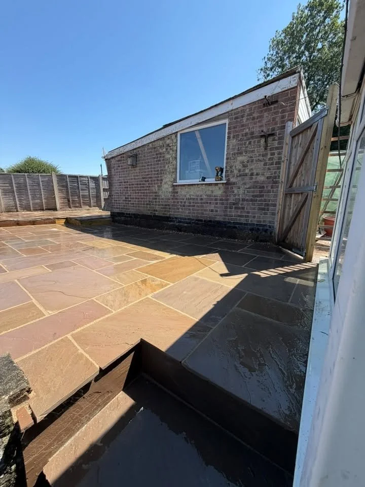 Newly paved patio with large stone tiles, partially wet from recent cleaning or sealing, next to a brick house with a window, a wooden fence, and a blue sky.