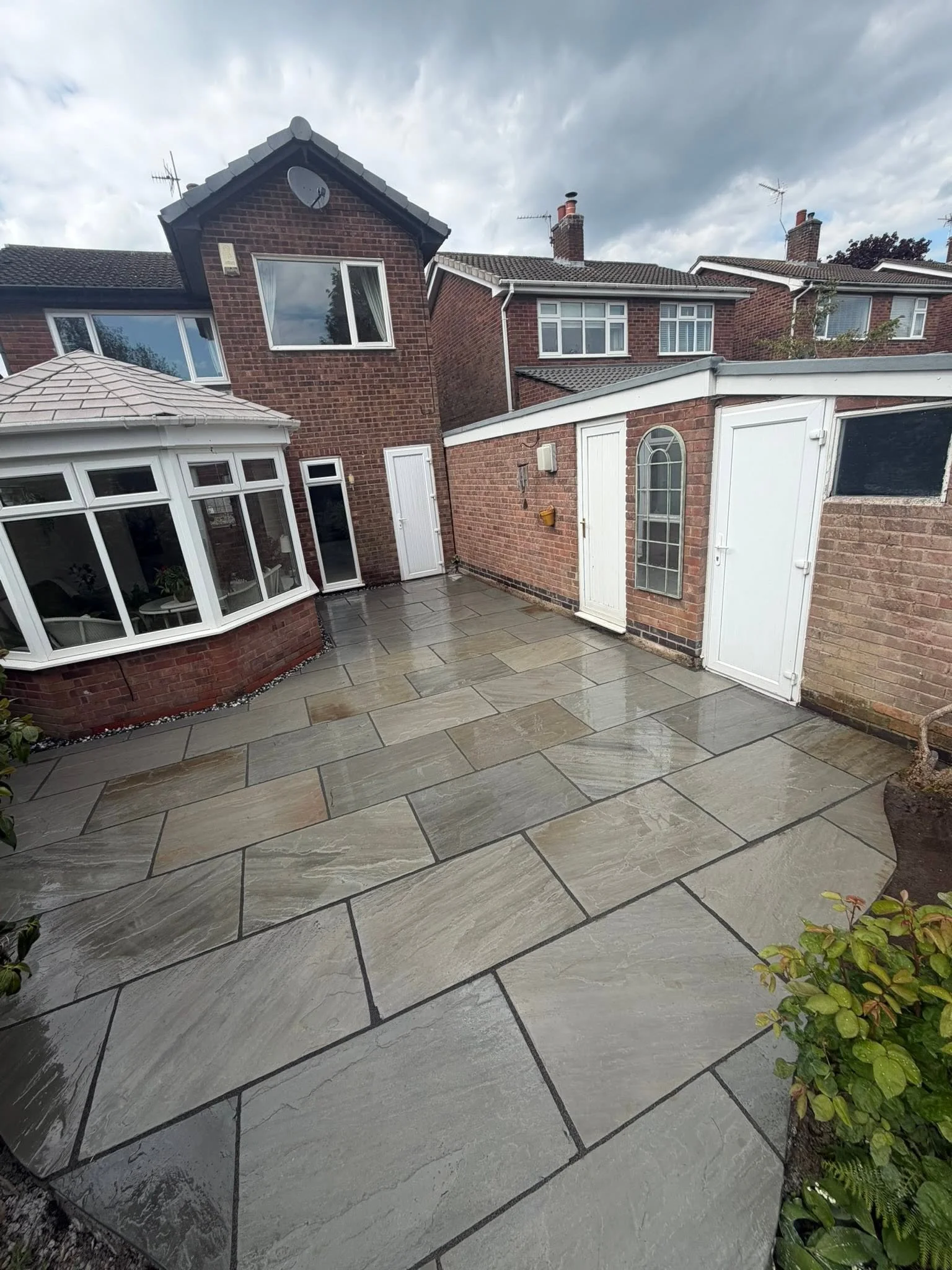 Wet outdoor patio with large grey stone tiles, adjacent to a brick house with white doors, windows, and a conservatory, with neighboring brick houses visible in the background under a cloudy sky.