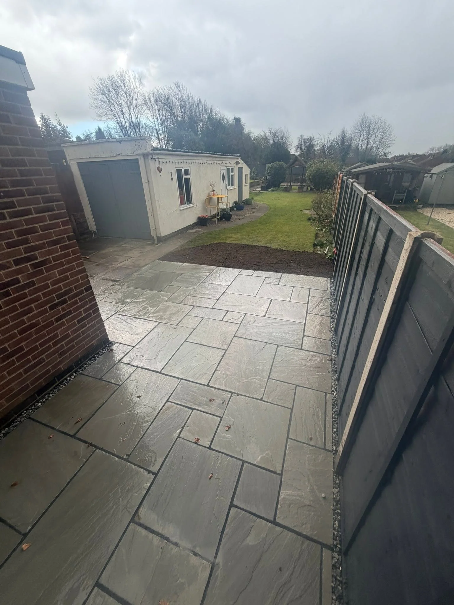 A backyard with a paved patio, a grassy area, and garden beds. There is a small white building with windows and a door, a greenhouse, trees, and bushes under a cloudy sky.