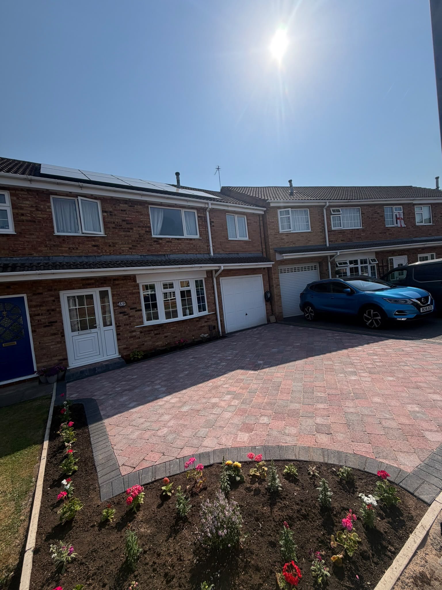 A brick townhouse with white-framed windows and garage doors, a driveway with a parked blue car, a front garden with pink and red flowers, and solar panels on the roof under a bright sunny sky.
