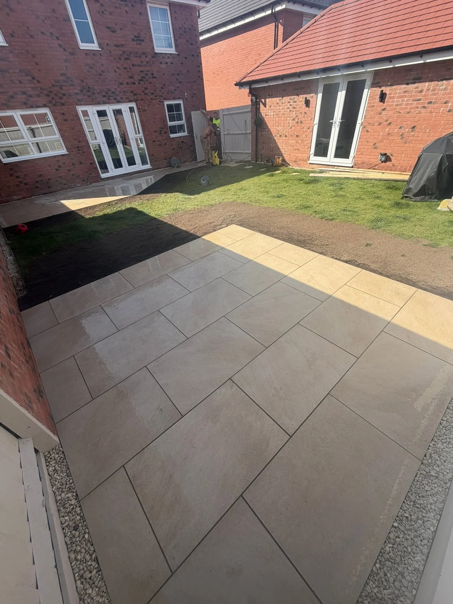 Backyard with new beige stone tile patio, green grass, brick house wall with French doors, and a person in a yellow vest working on the fence.