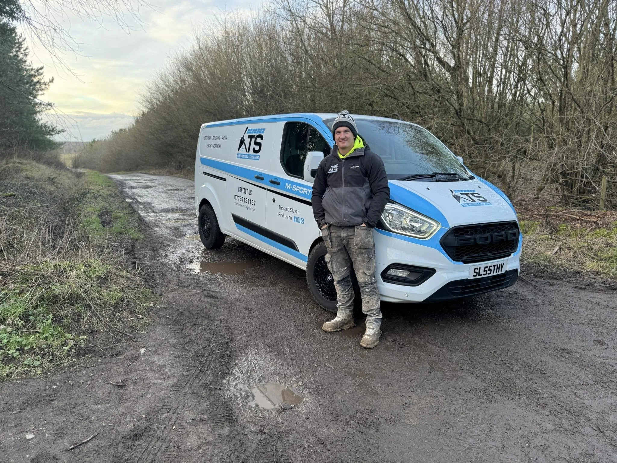 A man wearing a black jacket, gray pants, and a gray beanie with a neon yellow hoodie underneath, standing outdoors next to a white work van on a muddy, partially flooded dirt road with bare trees in the background.