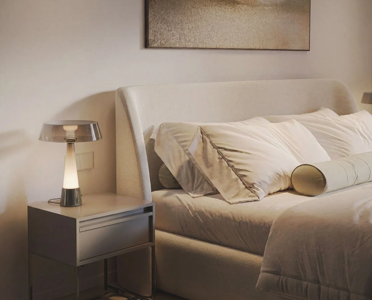 Letto matrimoniale con cuscini e copriletto beige, lampada da tavolo sul comodino accanto, quadro appeso sopra il letto, pareti di colore chiaro.