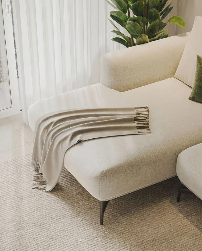Divano beige con un cuscino verde, una coperta beige stesa sul bracciolo e una pianta di grandi foglie verdi dietro.