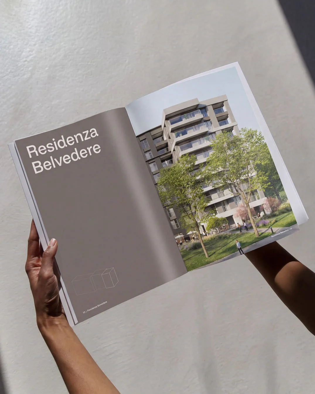 Una persona tiene tra le mani un opuscolo con l'immagine di un condominio moderno chiamato Residenza Belvedere, con molte piante e bambini che giocano nel parco