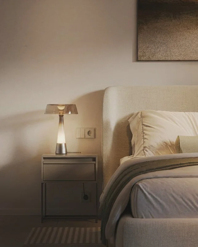 Letto con cuscini e una lampada da notte accesa su una  side table accanto a un muro.