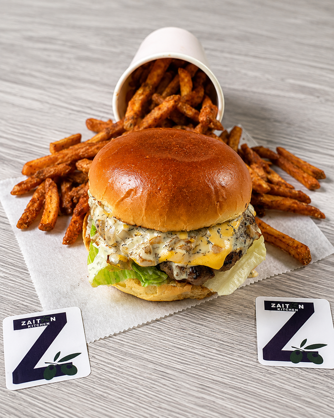 A cheeseburger with lettuce, melted cheese, and a sandwich bun, served with French fries in a paper cup on a gray wooden table.