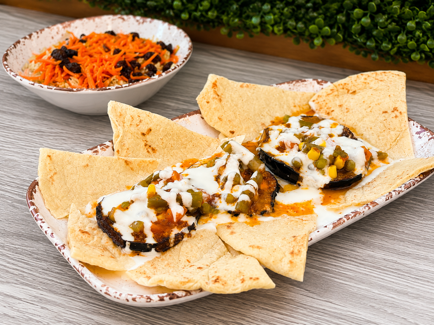 Plate of roasted eggplant topped with melted cheese, green peas, corn, and sauce, served with tortilla chips and a side of shredded carrot and black bean salad.