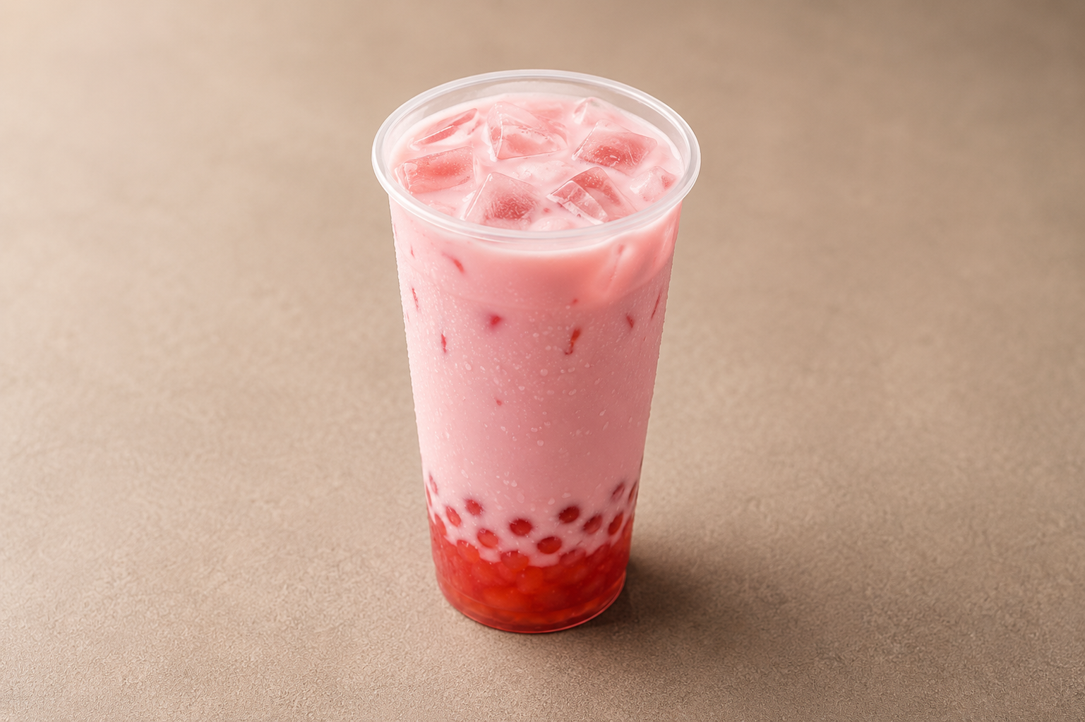 Pink bubble tea with tapioca pearls at the bottom and ice cubes on top, in a clear plastic cup on a beige surface.