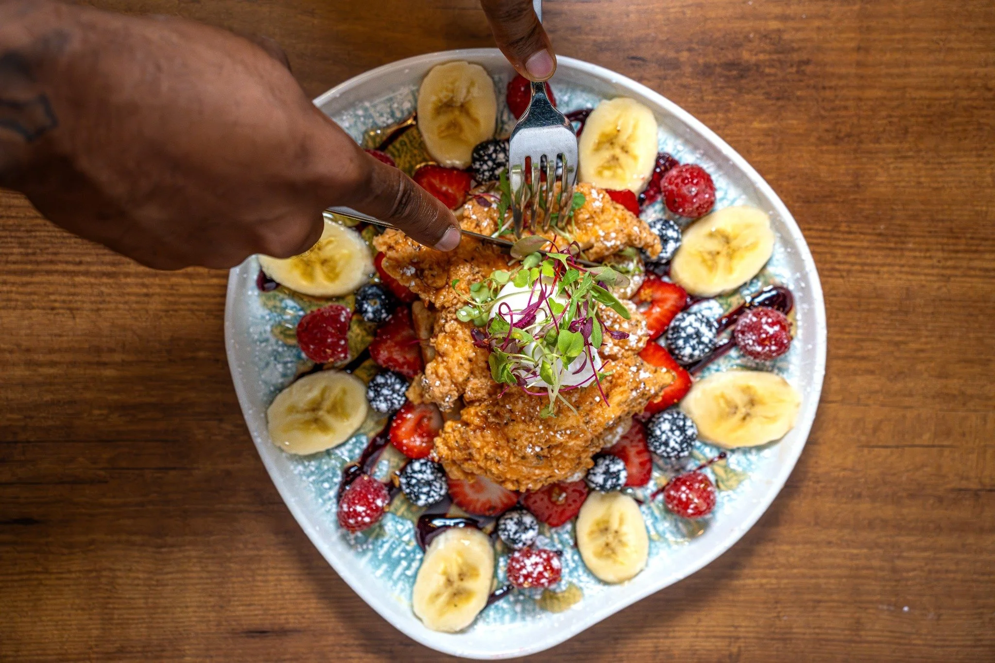 A decorative plate with sliced bananas, strawberries, blackberries, and blueberries, topped with microgreens and powdered sugar, with a piece of fried chicken or fish on top, drizzled with chocolate syrup, set on a wooden surface.