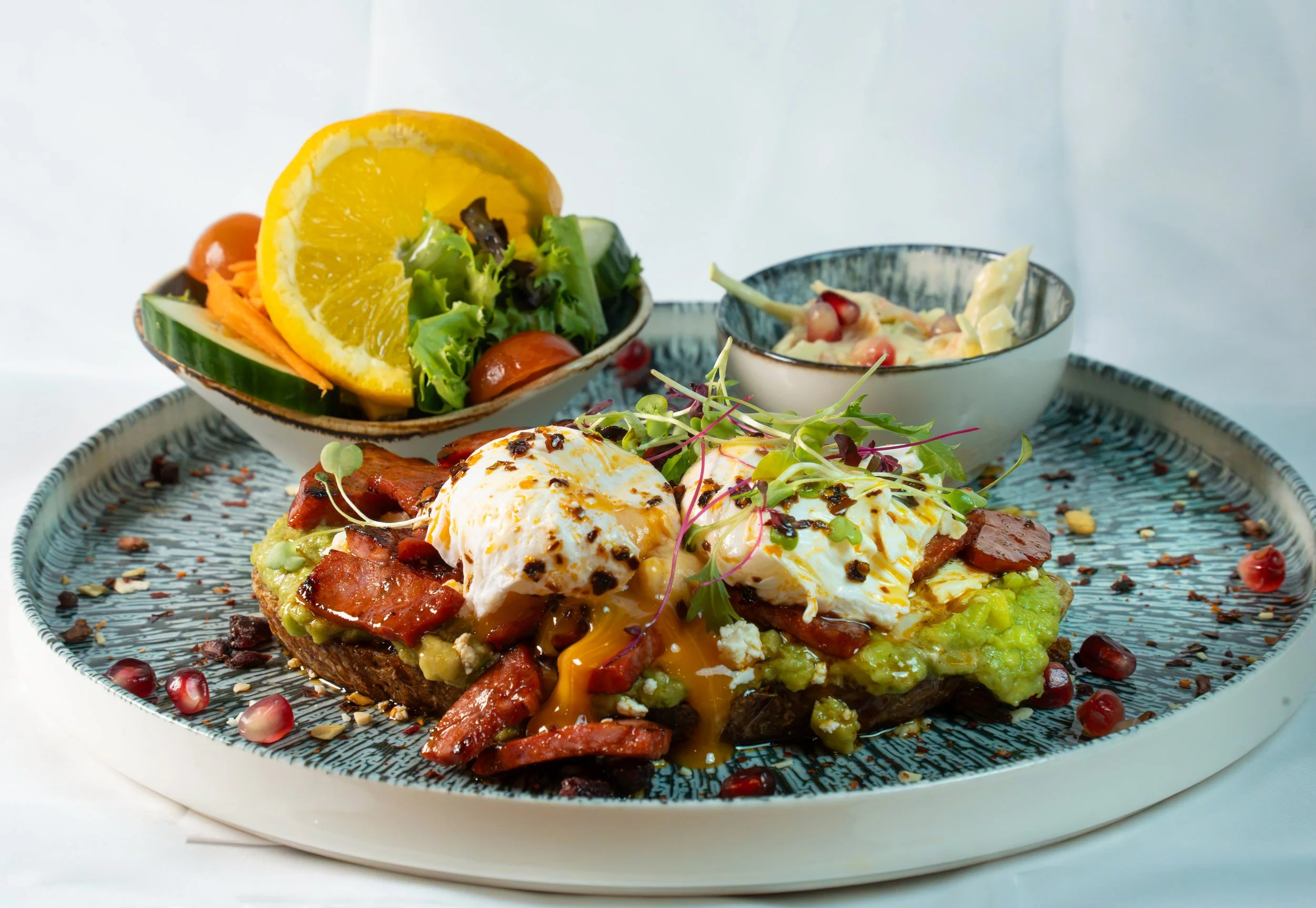 Close-up view of a dish with a poached egg on toast topped with bacon, microgreens, and sauce, served with a side salad and a small bowl of pasta salad on a patterned plate.
