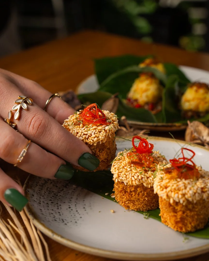Close-up of hand with green nail polish and rings reaching for a sesame-crusted appetizer garnished with red chili strands on a white plate.
