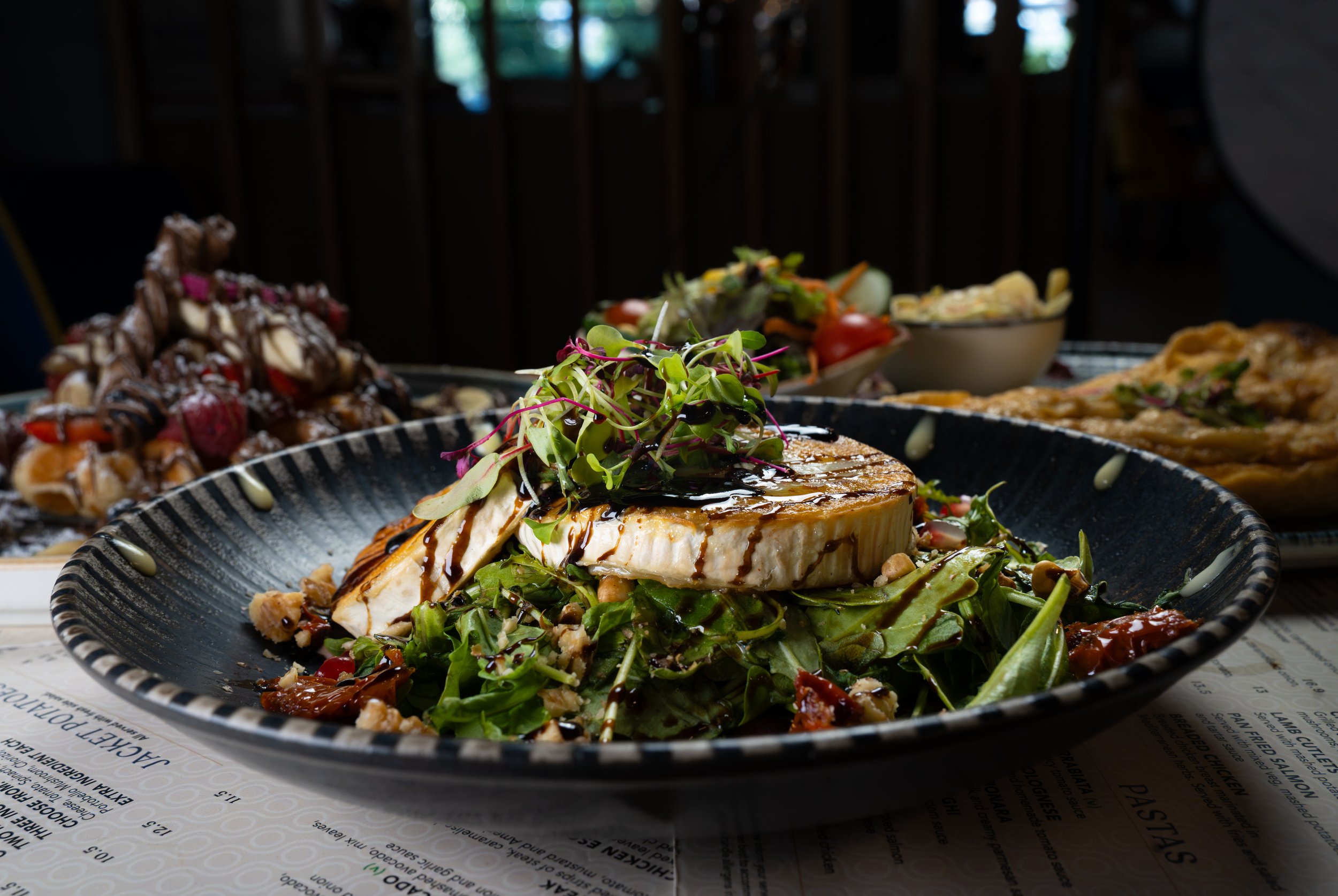A plate of grilled fish topped with microgreens, drizzled with balsamic glaze, served on a bed of salad with mixed greens and garnished with chopped nuts. In the background, there are other dishes, including pasta and a dessert, on a table with a dark background and natural light.