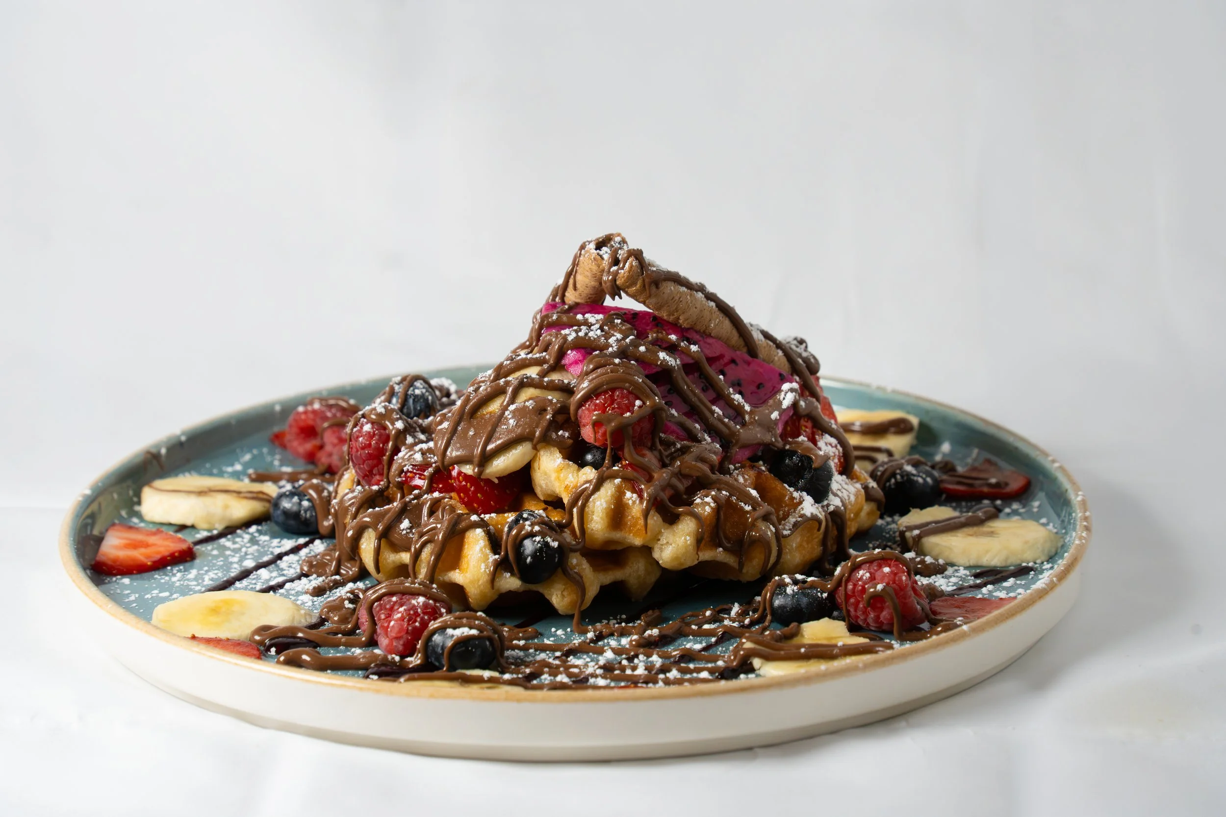 A plate of waffles topped with mixed berries, banana slices, chocolate drizzle, and powdered sugar.