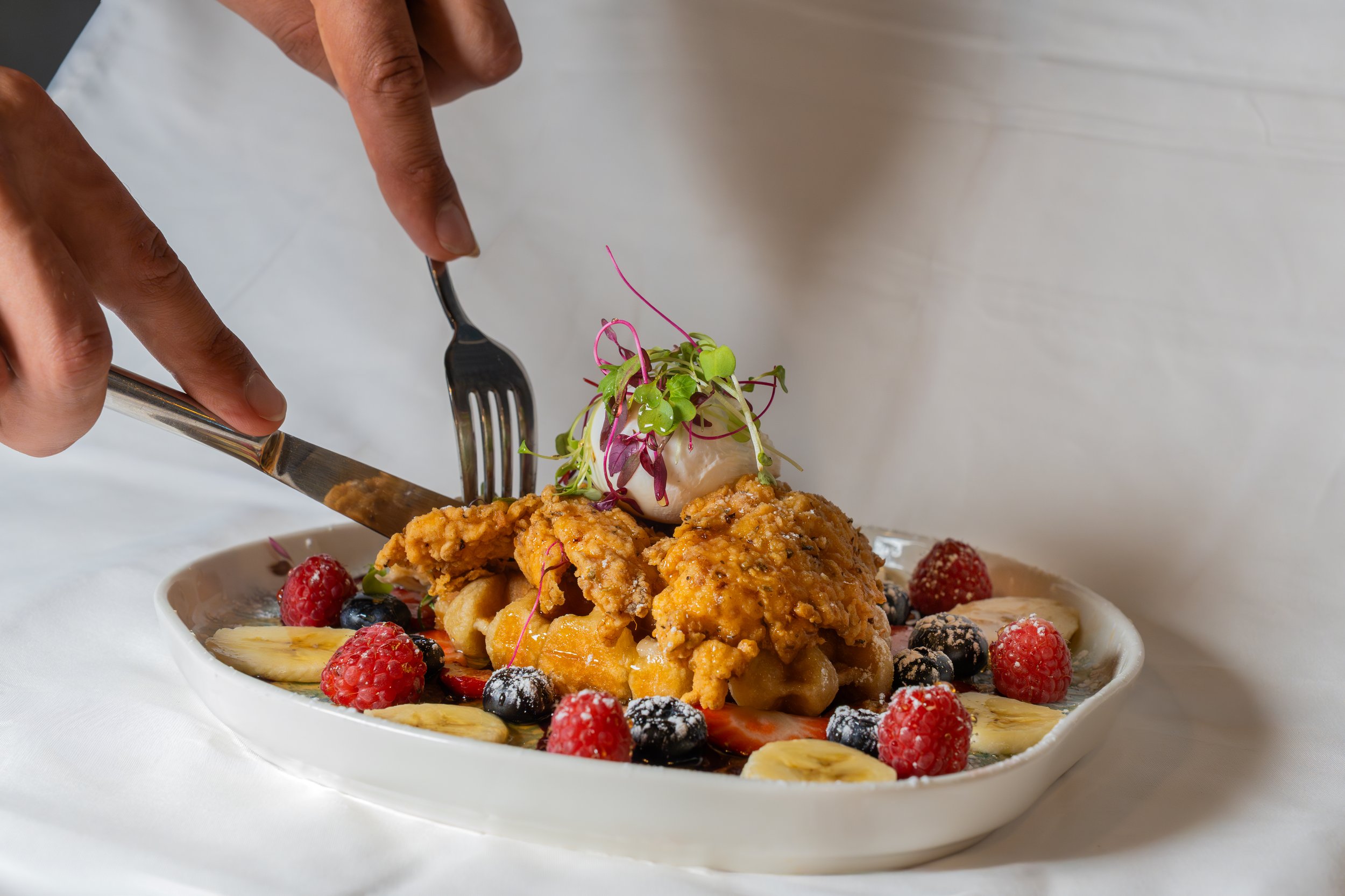 Close-up of a dessert plate with fried chicken, berries, banana slices, and microgreens, with a fork and knife.