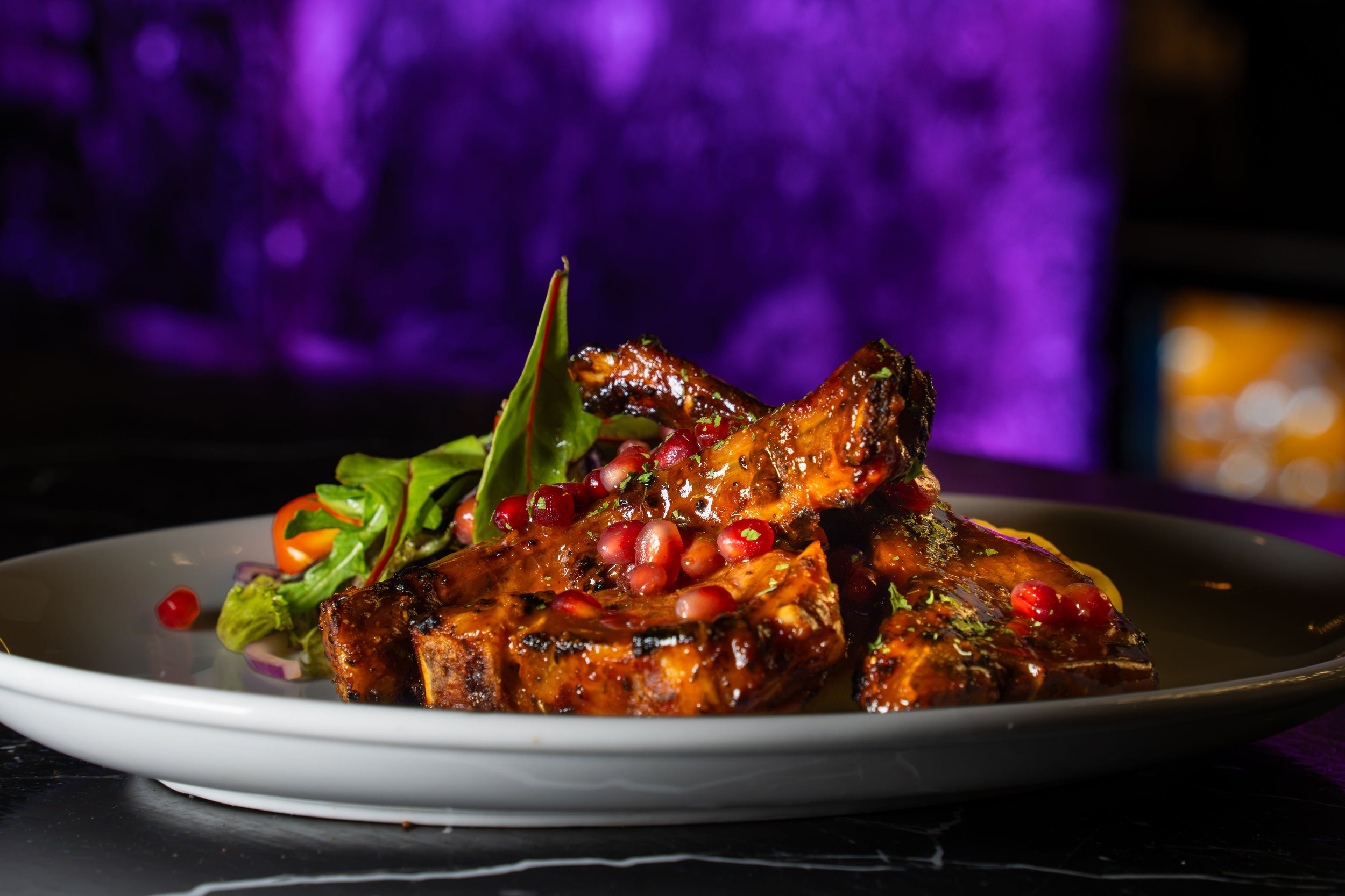 A plate of barbecued ribs topped with sauce and pomegranate seeds, garnished with fresh greens, against a blurred purple background.