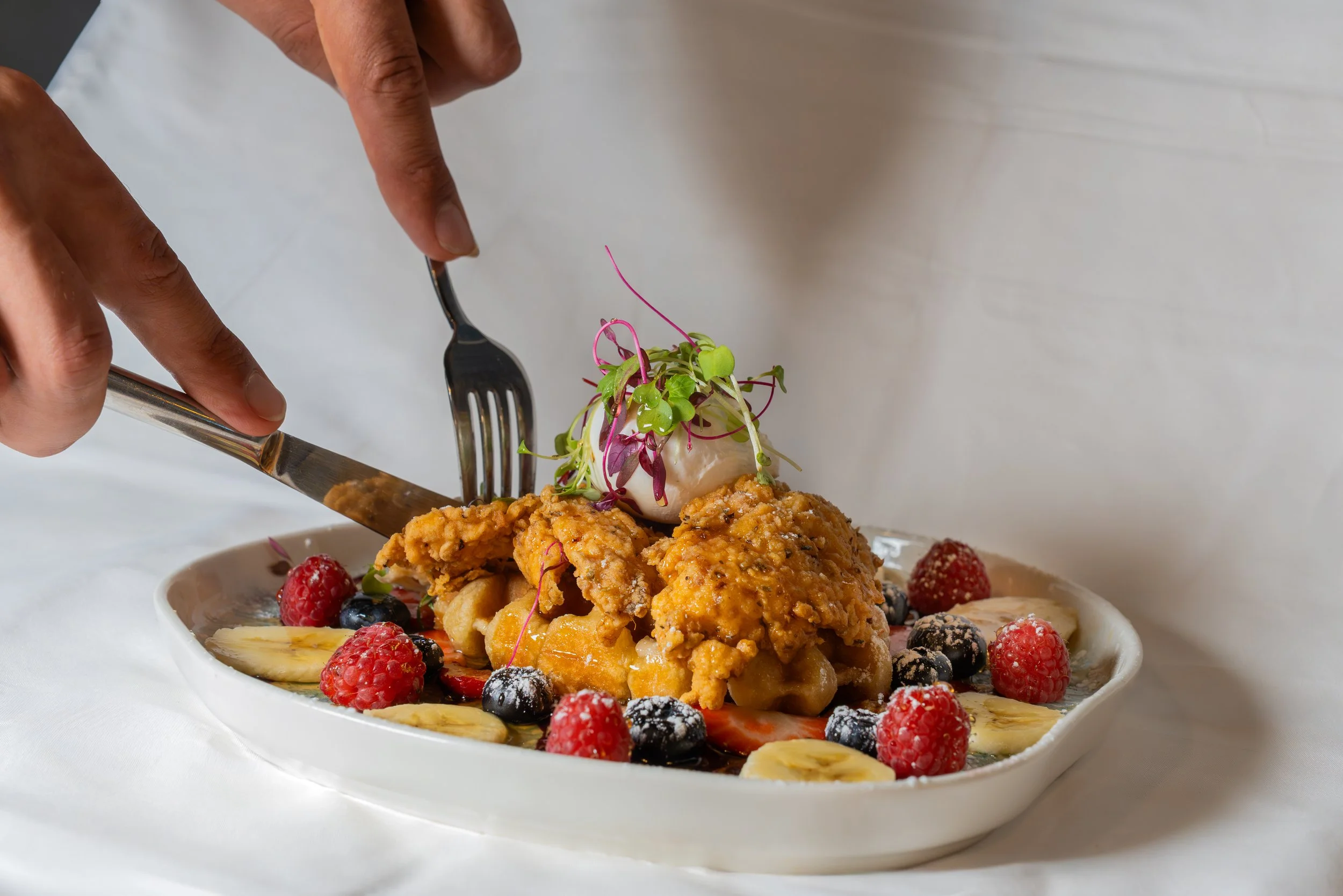 A plate of fried chicken topped with microgreens, surrounded by assorted berries and sliced bananas.