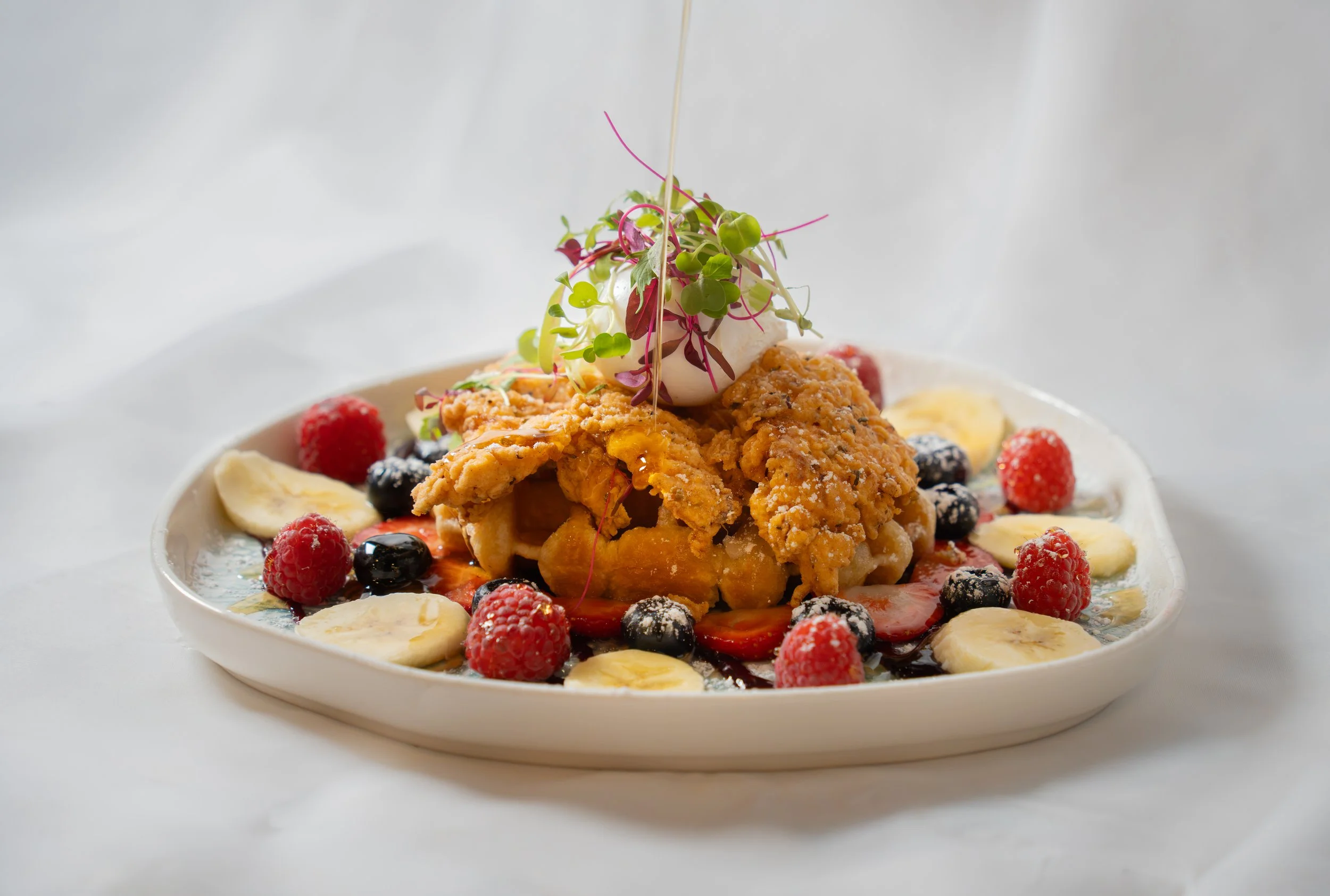 A dessert with fried dough, fresh raspberries, blueberries, sliced bananas, and a dollop of whipped cream topped with microgreens in a white bowl.